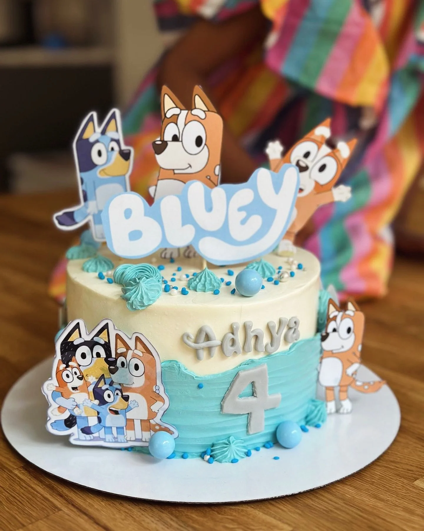 A two-tiered birthday cake decorated with characters from the animated series Bluey. The cake has white and blue frosting with blue icing swirls and spheres. Cartoon character cutouts, including Bluey, Bingo, and Bandit, are on top and around the cak