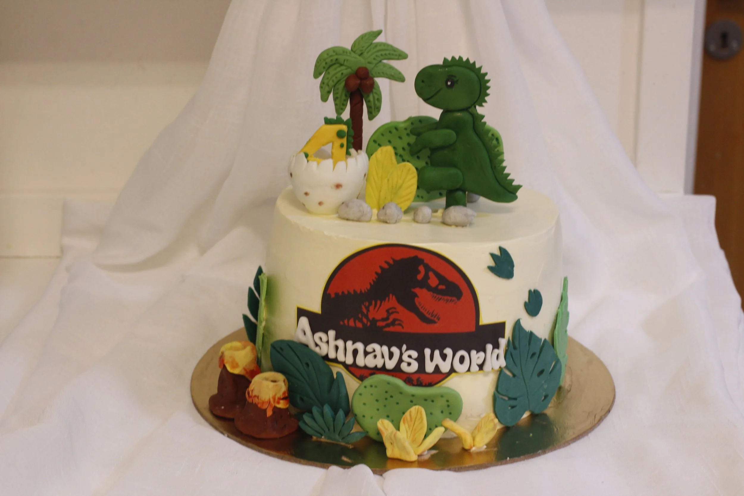 A themed birthday cake with jungle and dinosaur decorations, featuring the logo from Jurassic Park and the text 'Ashnavi's World', topped with edible decorations of a dinosaur and jungle elements.