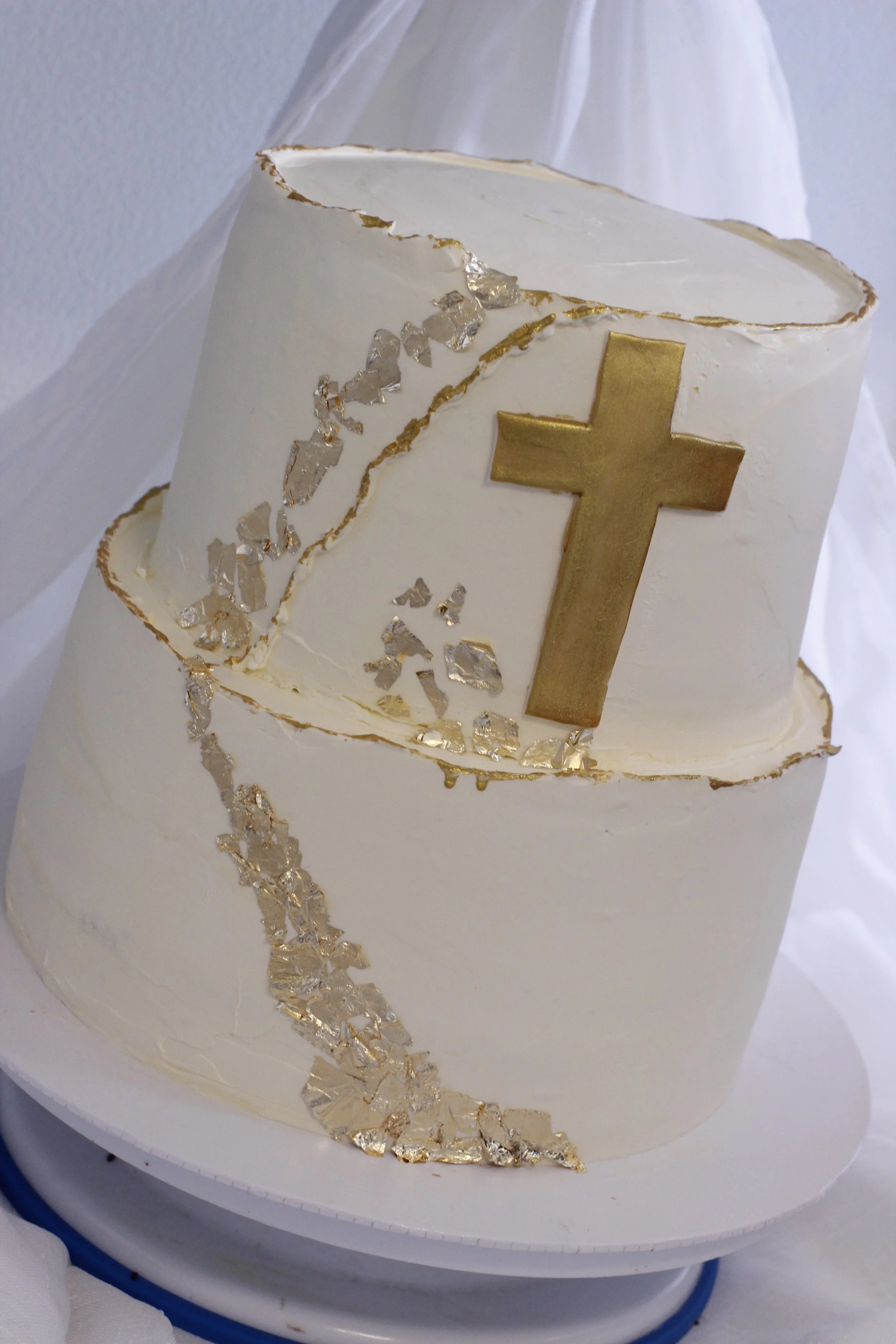 A two-tiered white cake decorated with a gold cross, gold and silver accents, and artistic gold leaf details, celebrating a religious occasion.