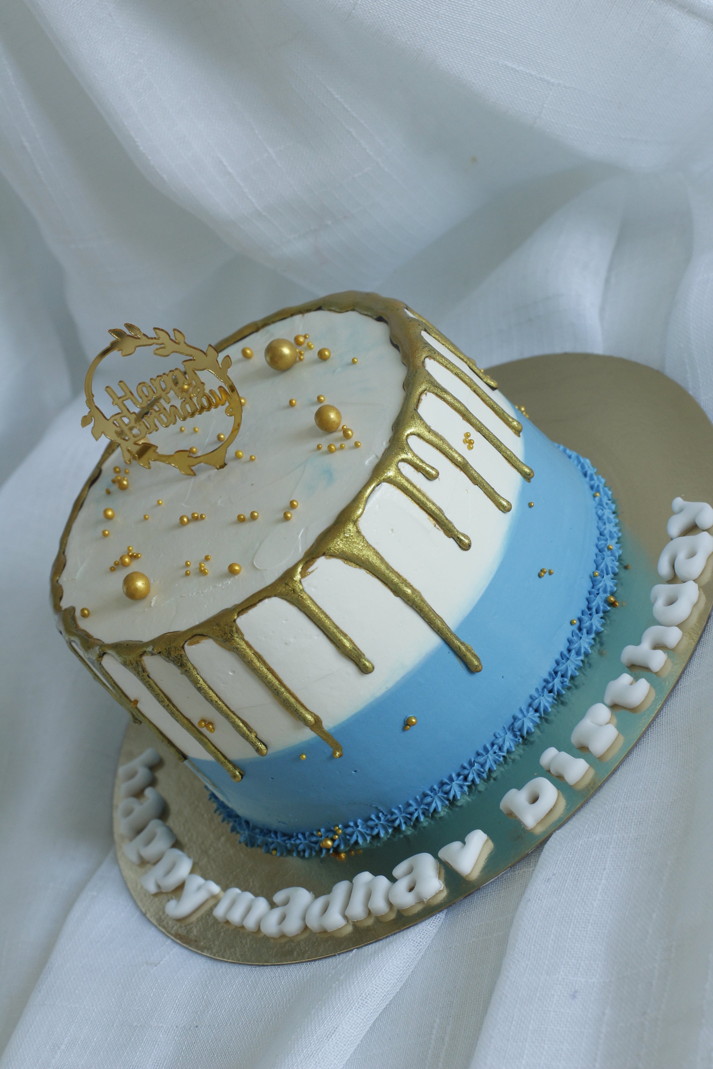 A round birthday cake with white and blue icing, decorated with gold drips and gold balls, topped with a 