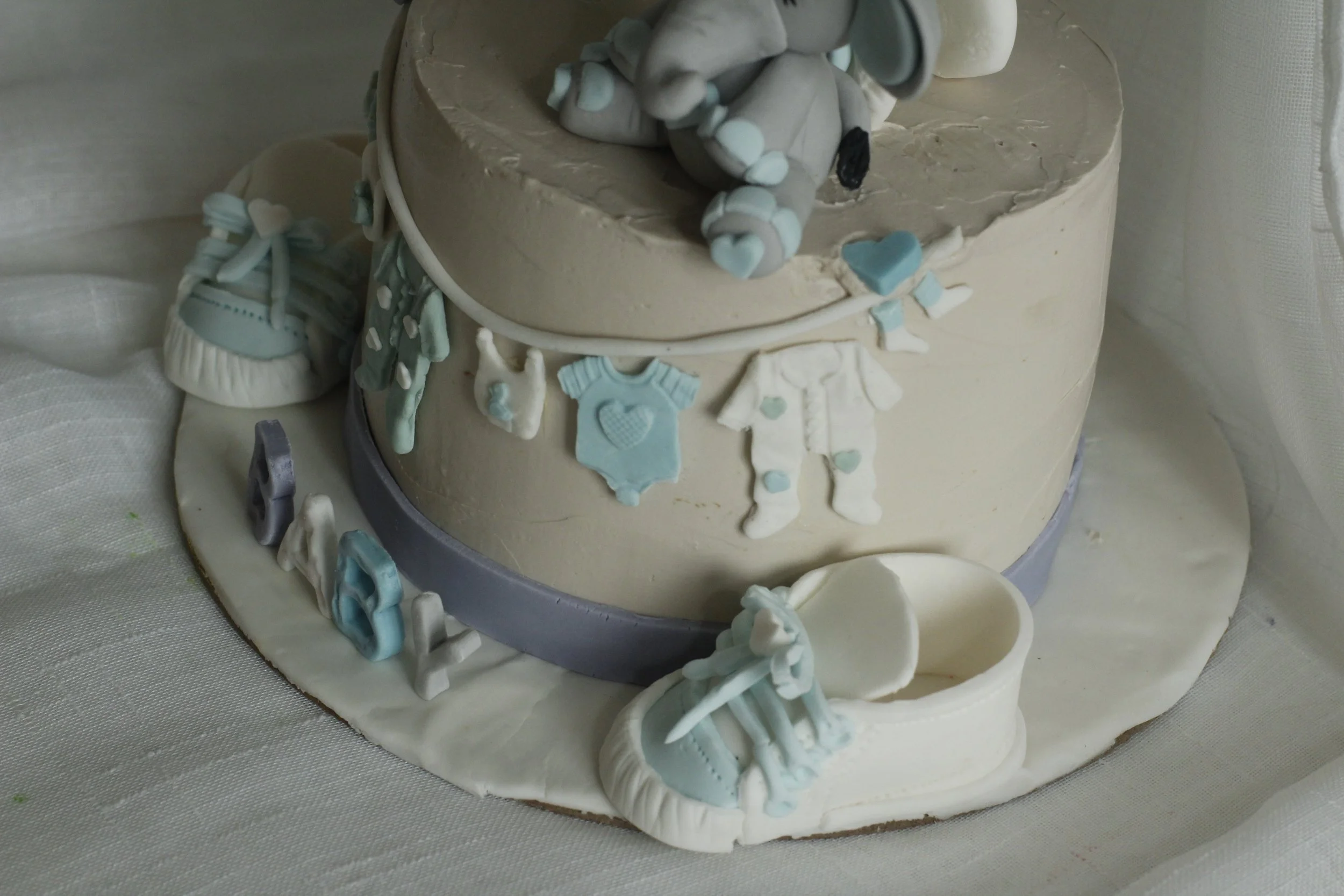 A baby shower cake decorated with baby clothes and shoes in soft pastel colors, featuring a stuffed elephant on top.
