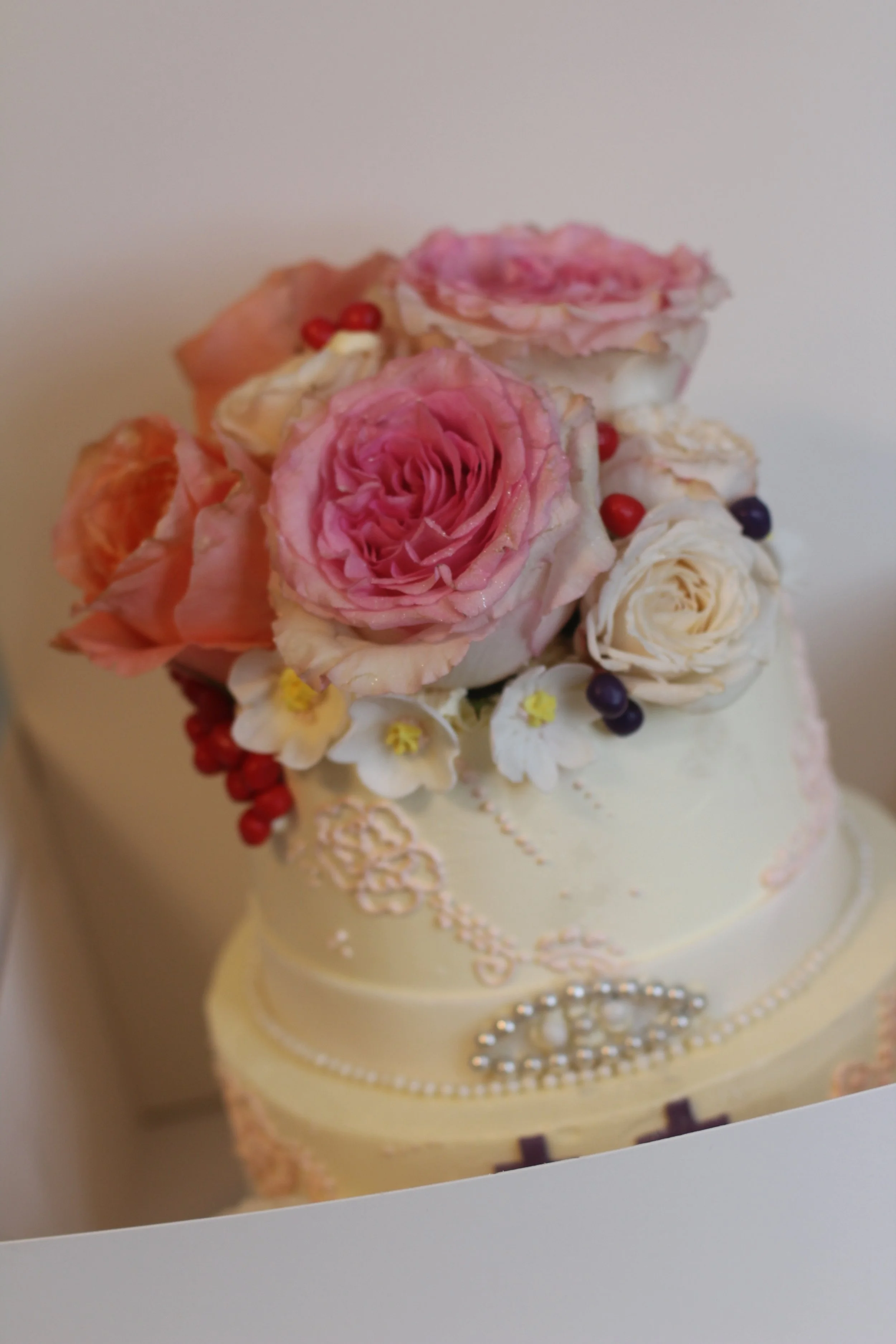 Cake decorated with pink, white, and peach roses, small white flowers, colorful berries, and pearl-like embellishments.