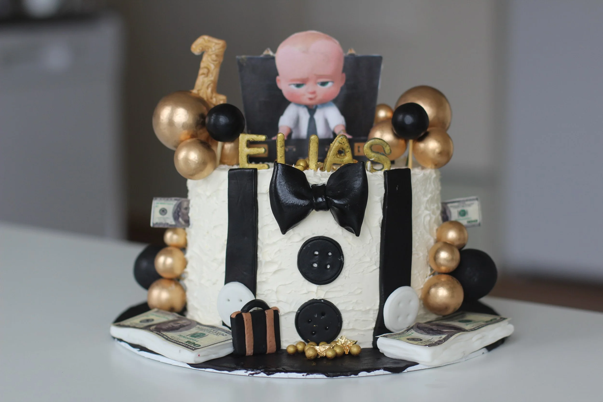 Decorative birthday cake with a money theme, featuring black, white, and gold accents, a black bow tie, buttons, a photograph of a boy, golden and black ball decorations, gold 'ELIAS' letters, a gold '1' topper, and dollar bills around the base.