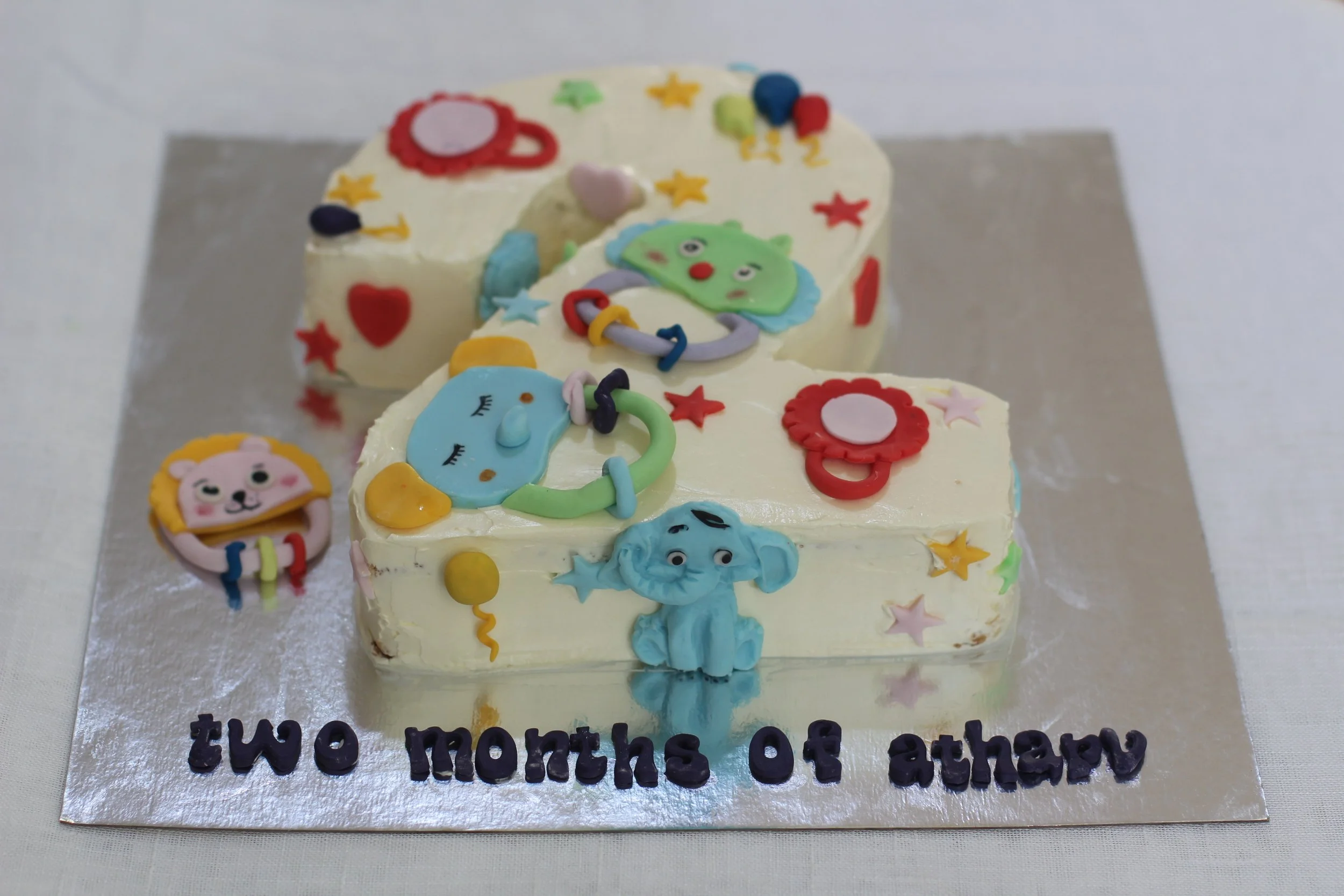 A birthday cake in the shape of the number two, decorated with colorful baby-themed fondant animals and toys, with the words 'two months of athar' written on a silver board at the base.