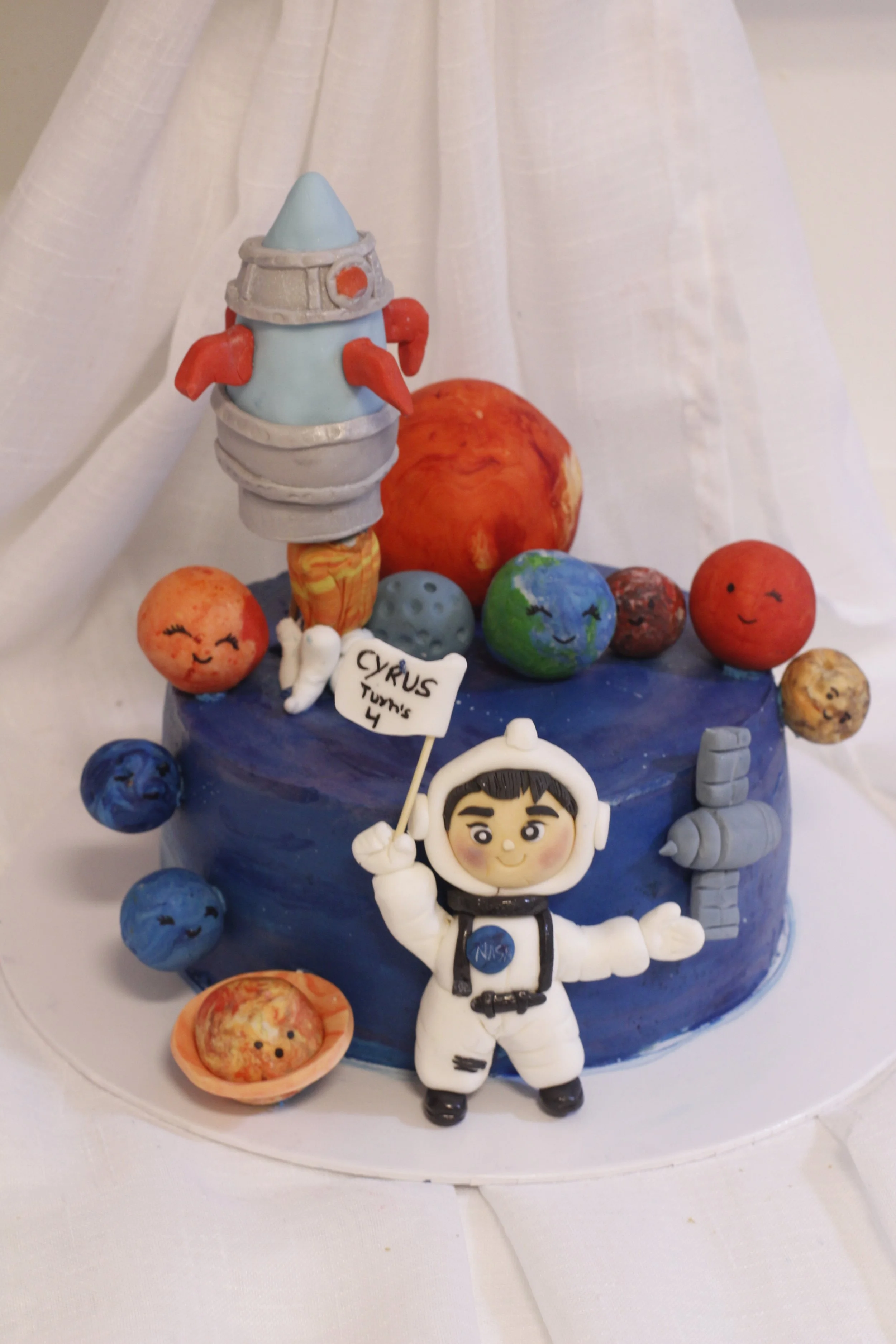 A space-themed cake decorated with planets, a rocket, an astronaut figurine, and a sign that says "Cyrus Turns 4."