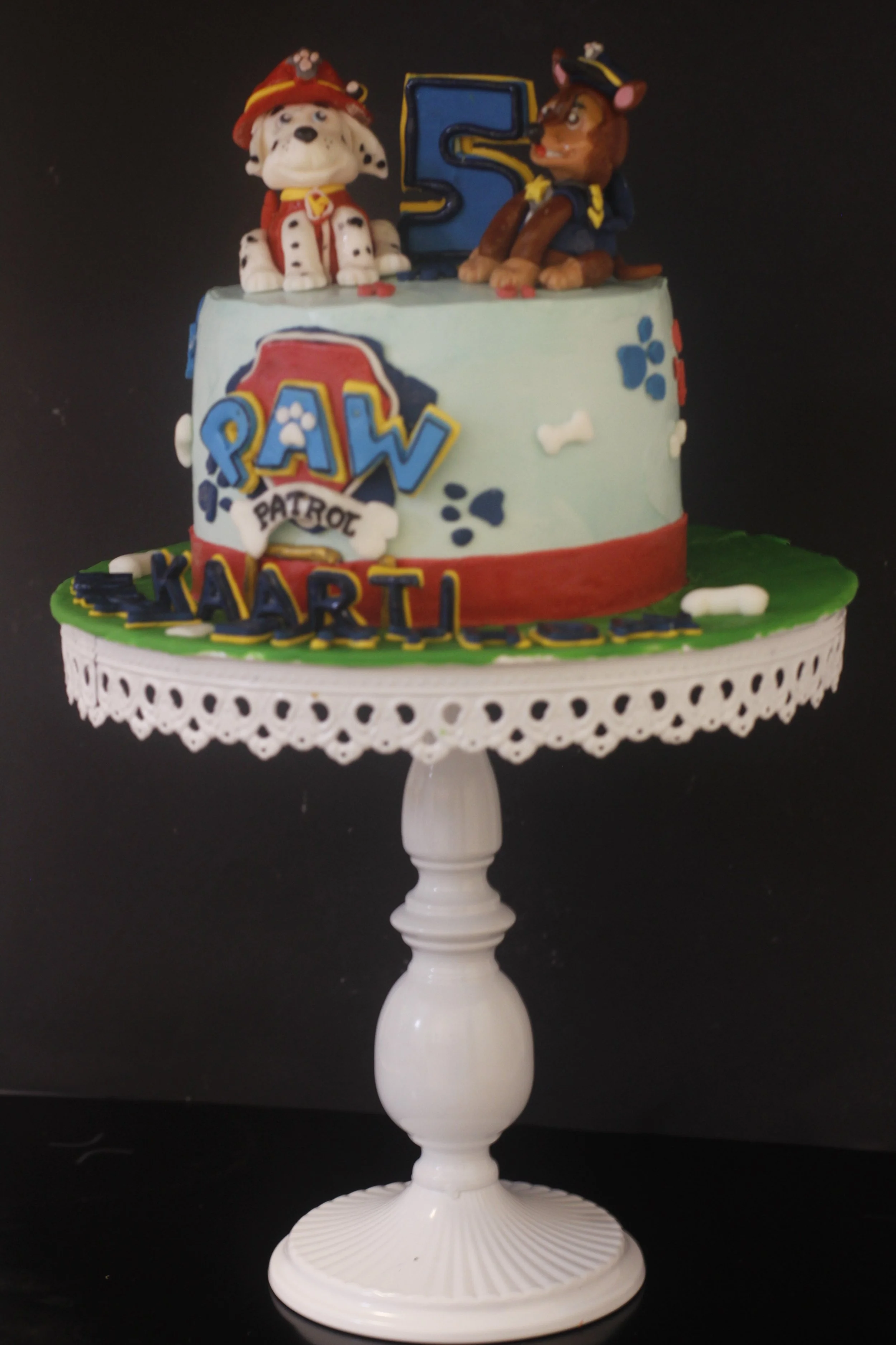 A children's birthday cake themed from the children's show "Paw Patrol," featuring figurines of dogs, a number five candle, and the child's name "Kartik" on the cake.