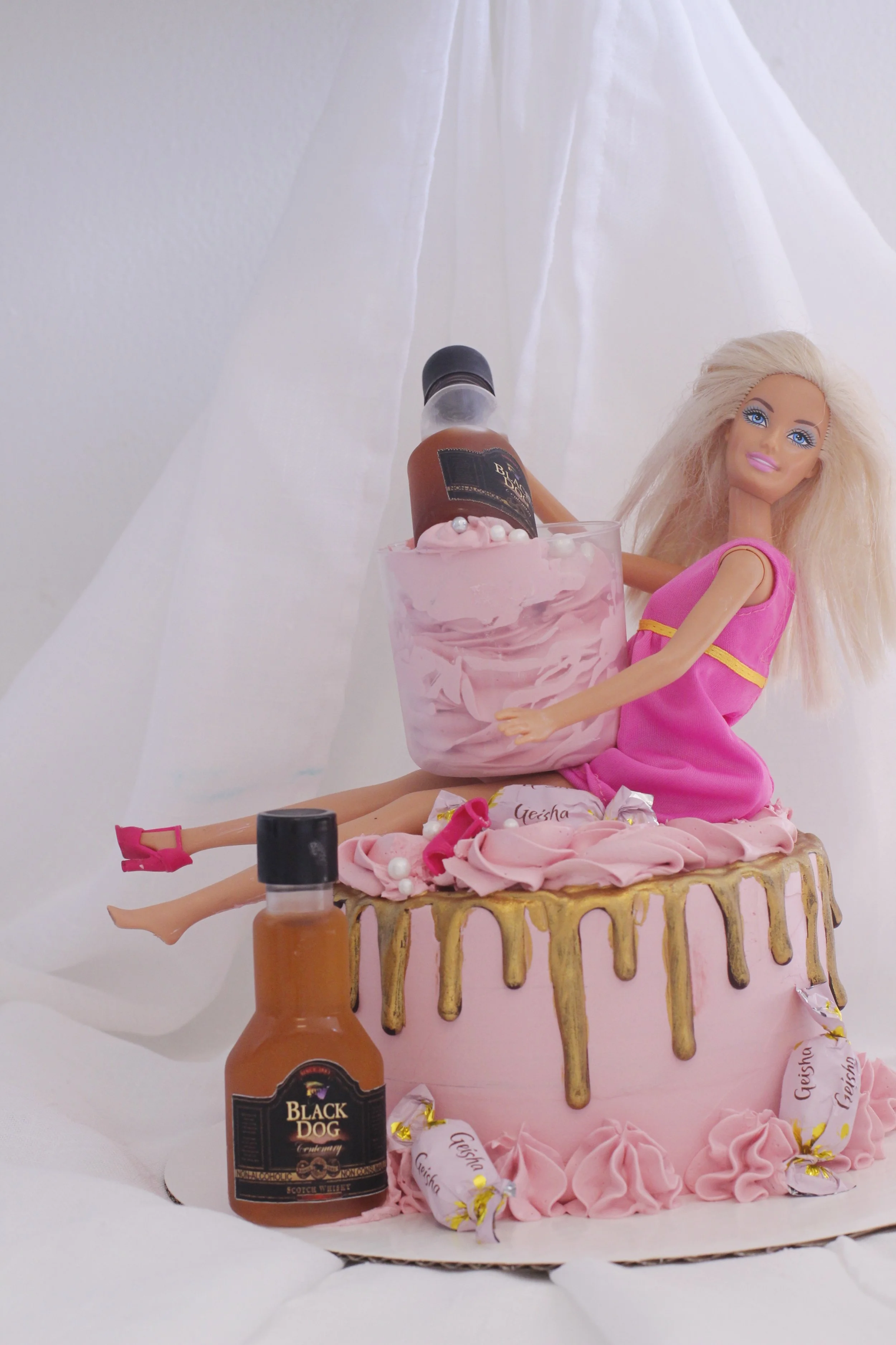 A Barbie doll sitting on a pink cake with gold accents, surrounded by small bottles resembling alcohol and candy, with a white fabric background.