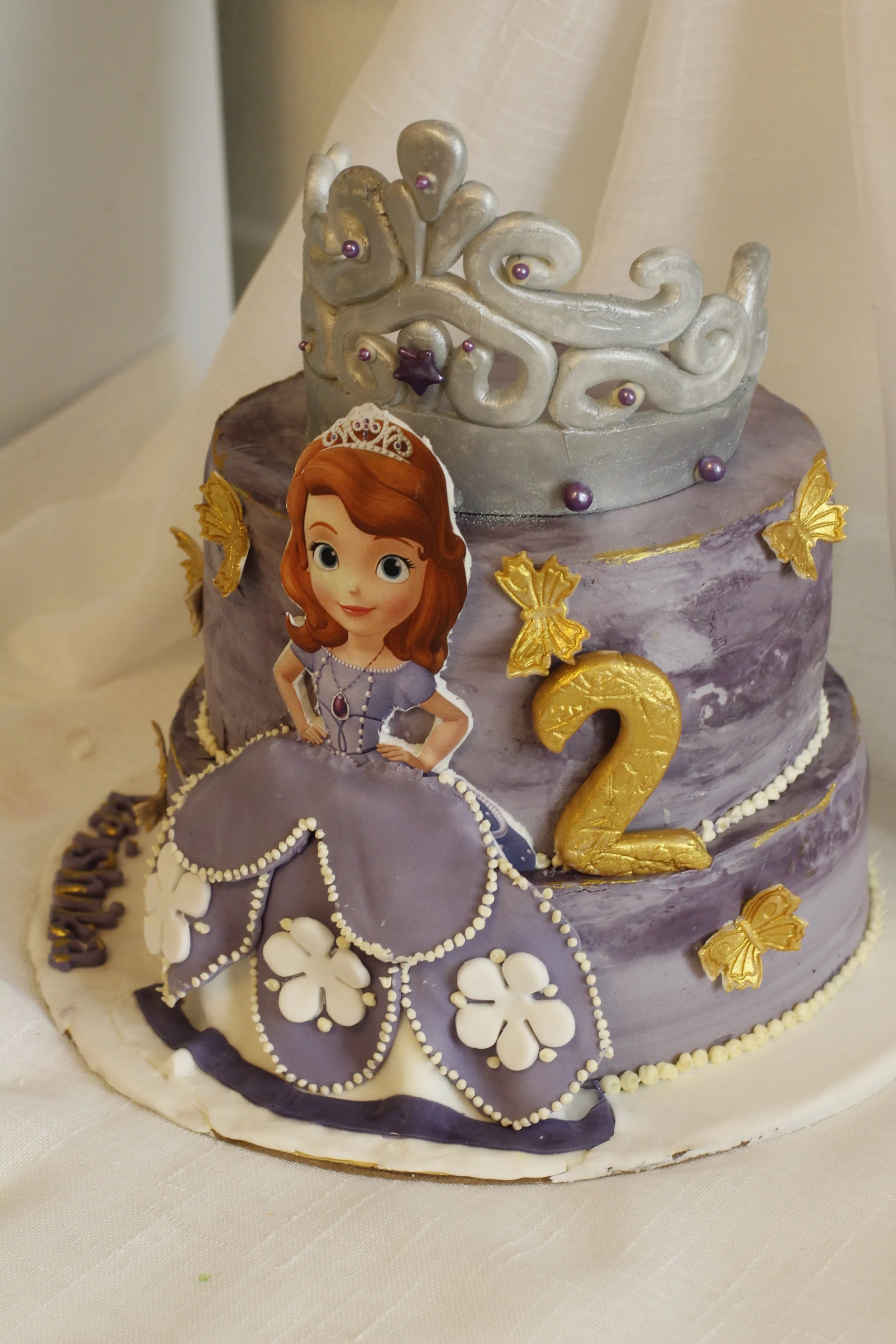 A purple, three-tiered birthday cake decorated with a princess figurine, a silver tiara on top, gold butterfly and number 2 decorations, and a paper cutout of a princess character.