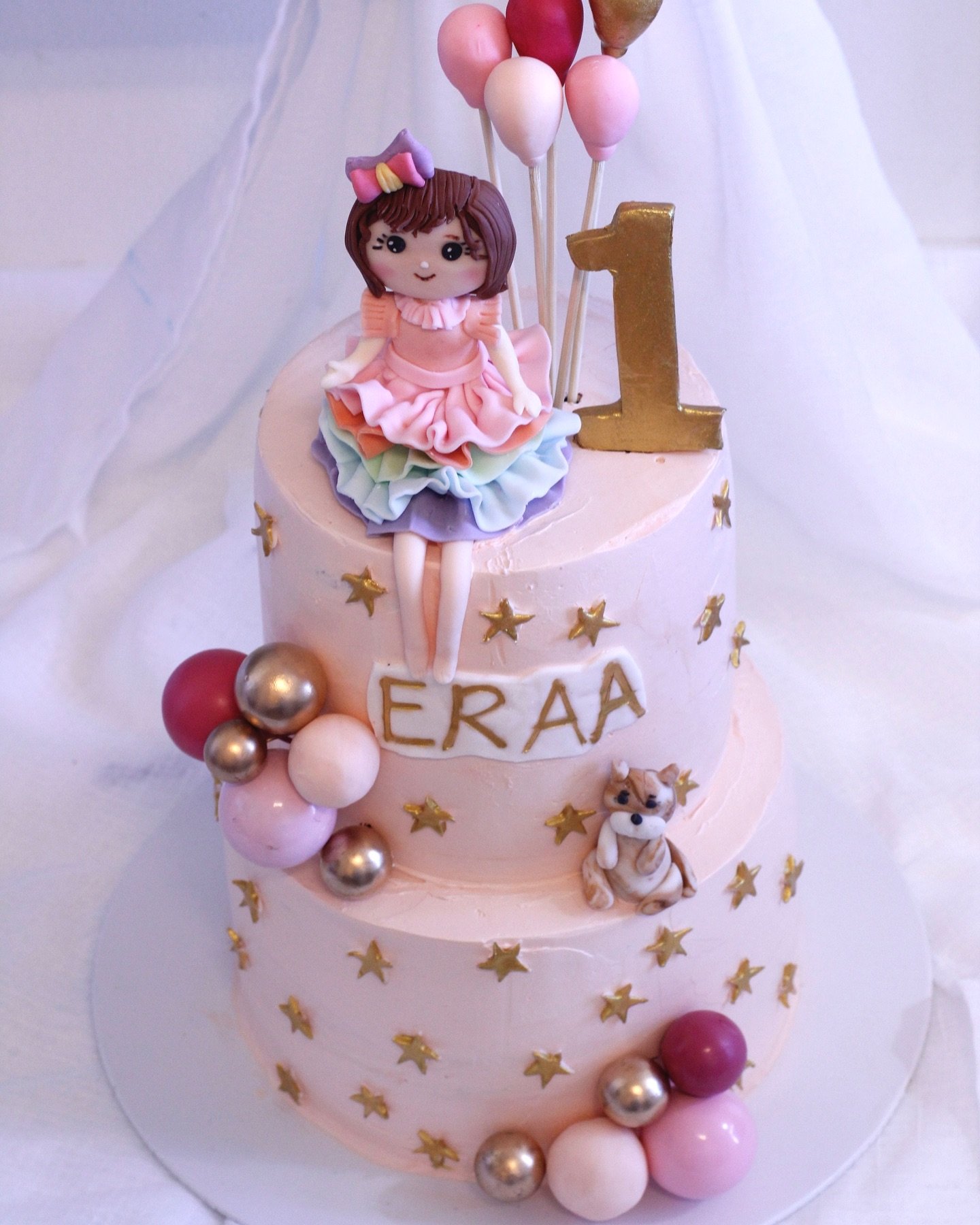 A two-tier pink birthday cake decorated with gold stars, pink, purple, white, and silver spheres, and a fondant girl doll sitting on top. The doll has brown hair, a purple bow, a pastel rainbow ruffled dress, and is sitting with legs dangling. There 