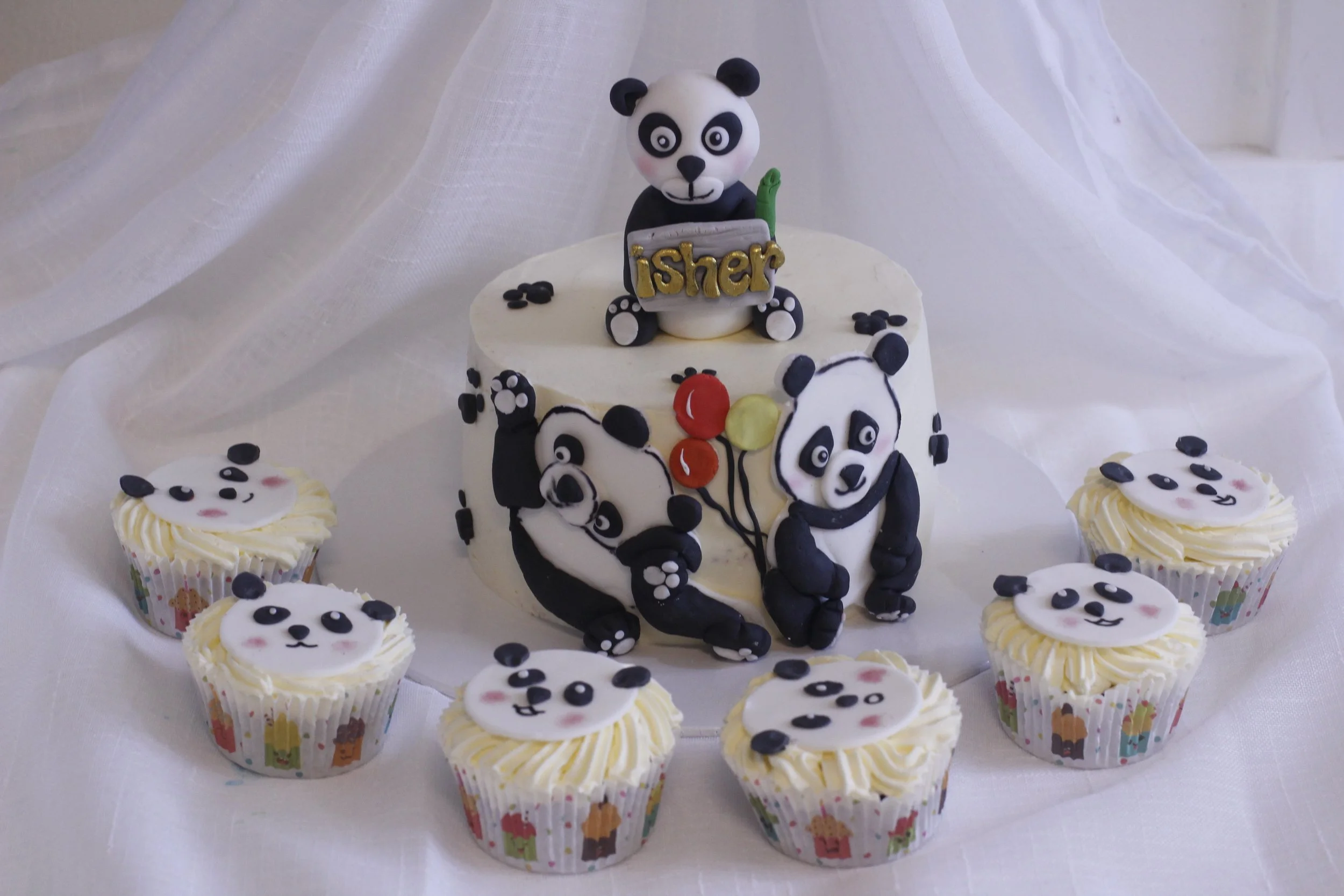 A birthday cake with panda bear decorations and cupcakes, featuring panda faces on the cupcakes, a panda holding a sign that says "Isher" on top, and panda and balloon designs on the cake.