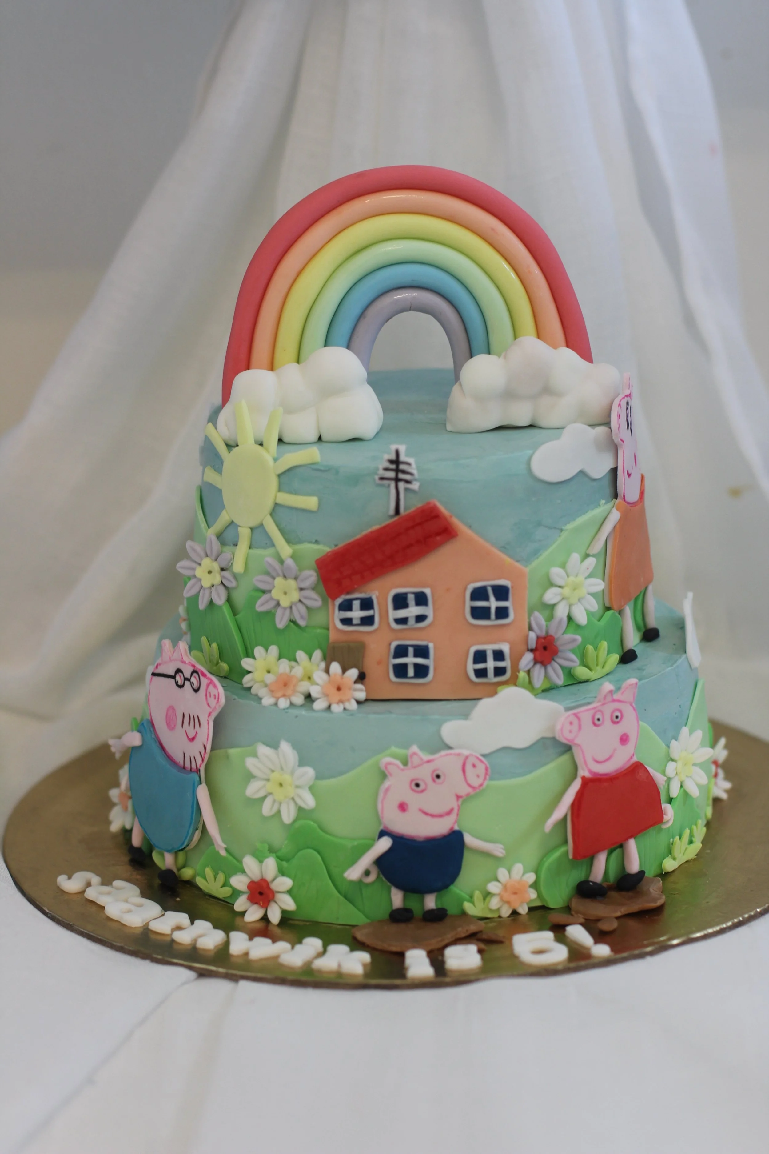 Colorful cake decorated with Peppa Pig characters, flowers, a rainbow, sun, house, and clouds.