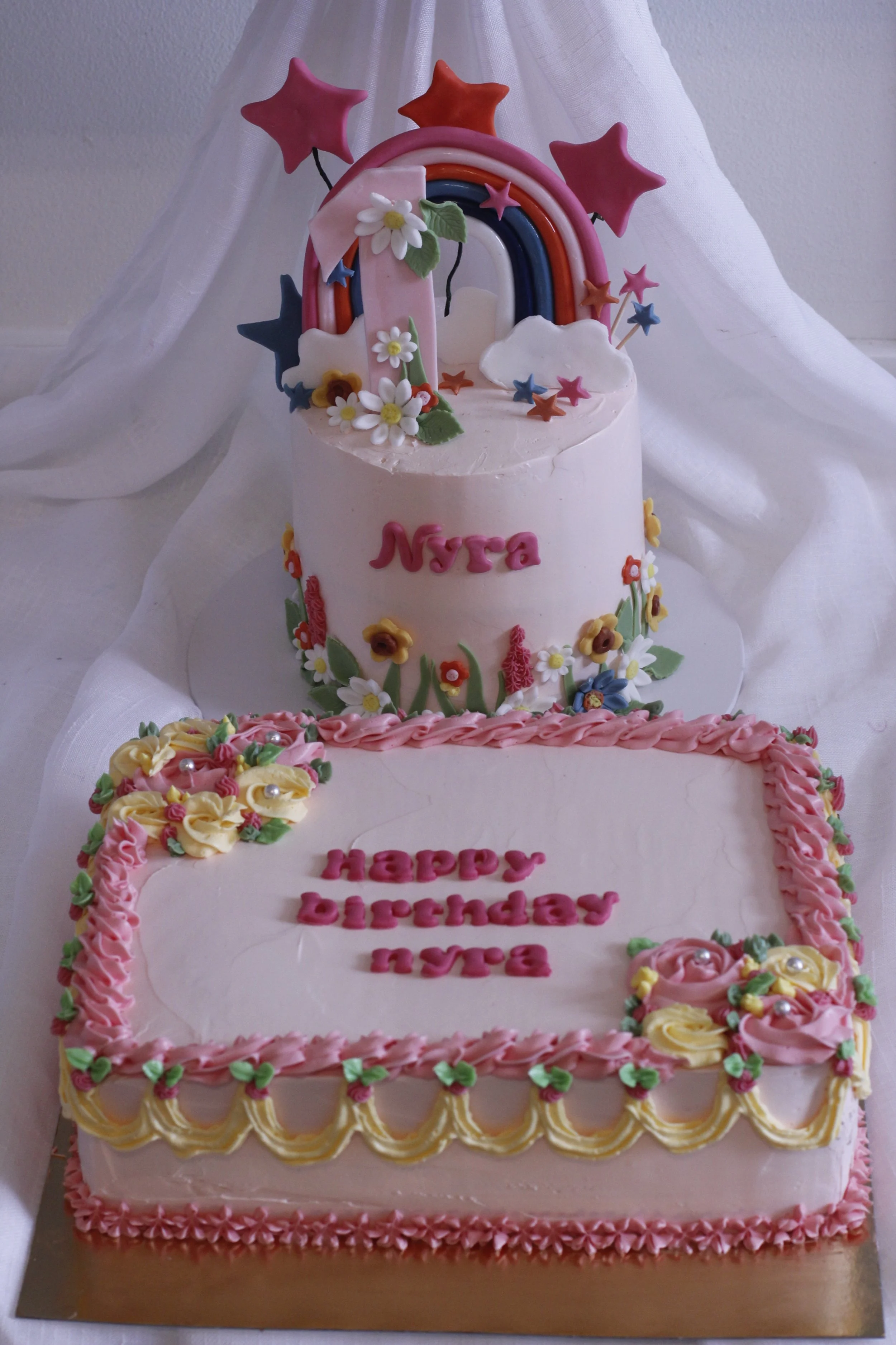 Pink birthday cake with colorful stars, a rainbow, flowers, and the message 'Happy Birthday Nyra' on it.