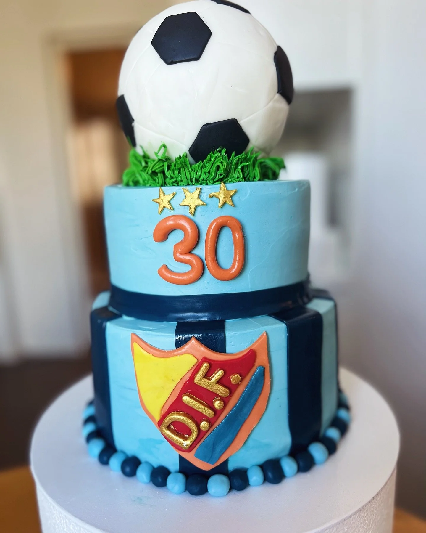 Decorative birthday cake with a soccer theme, featuring a soccer ball on top, the number 30, and a shield emblem with the word DIOK, in blue, orange, yellow, and red colors.