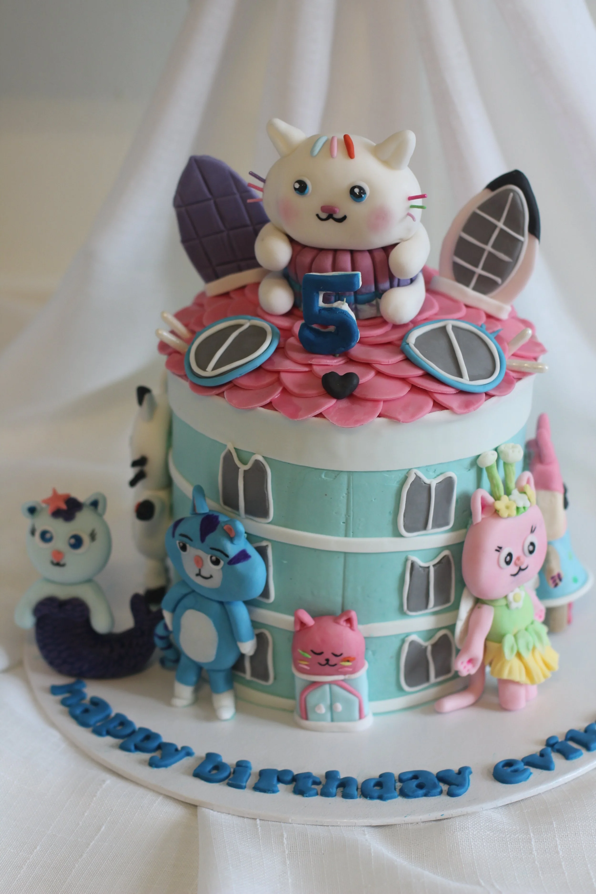 Colorful birthday cake decorated with cartoon cat characters and a blue number 5 candle on top, with a white cat sitting on the cake, surrounded by various playful cat figures.