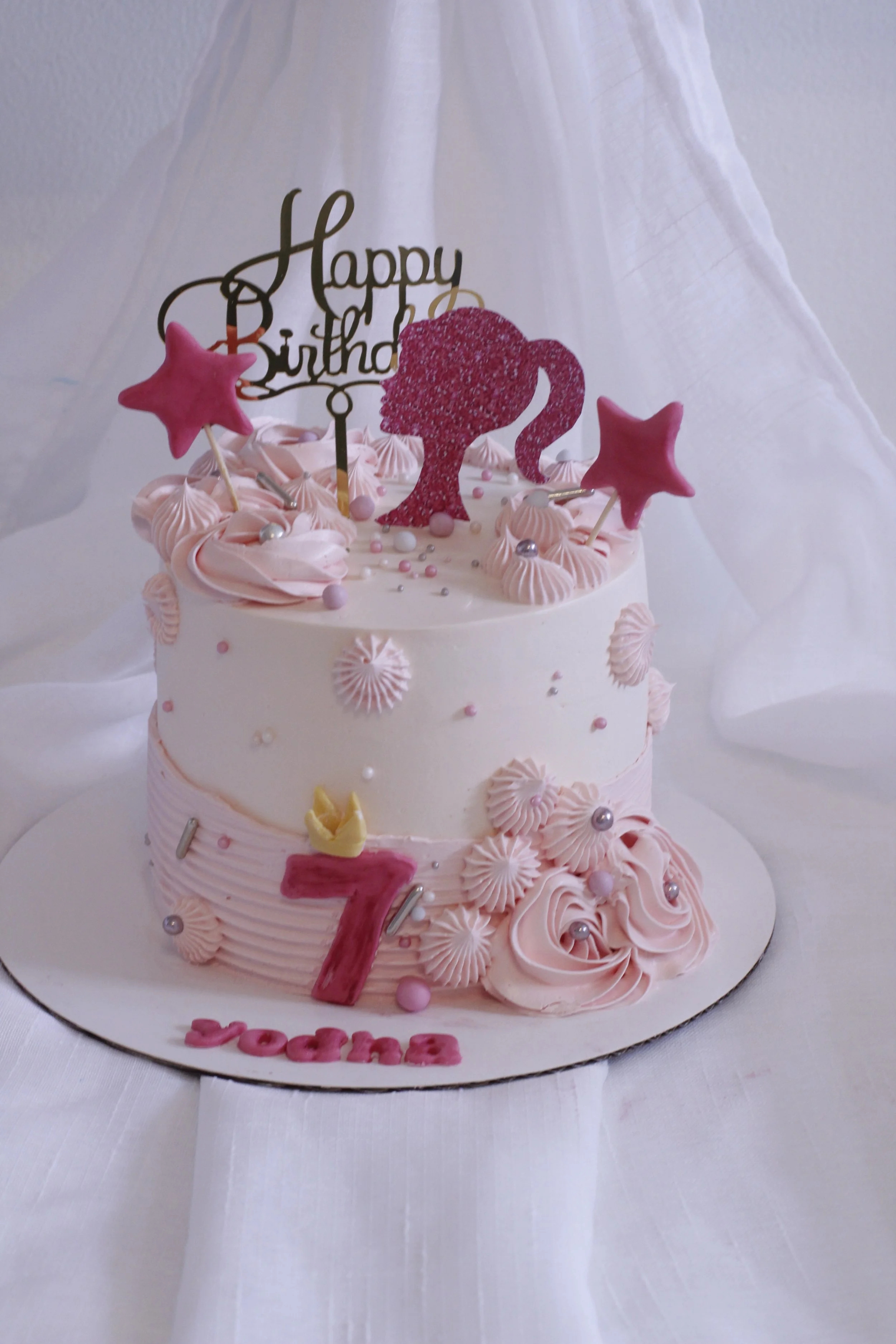 Pink and white birthday cake with star and flamingo decorations, a 'Happy Birthday' topper, and a large pink number 7 with the name 'Yorre' below, on a white cloth-covered surface.
