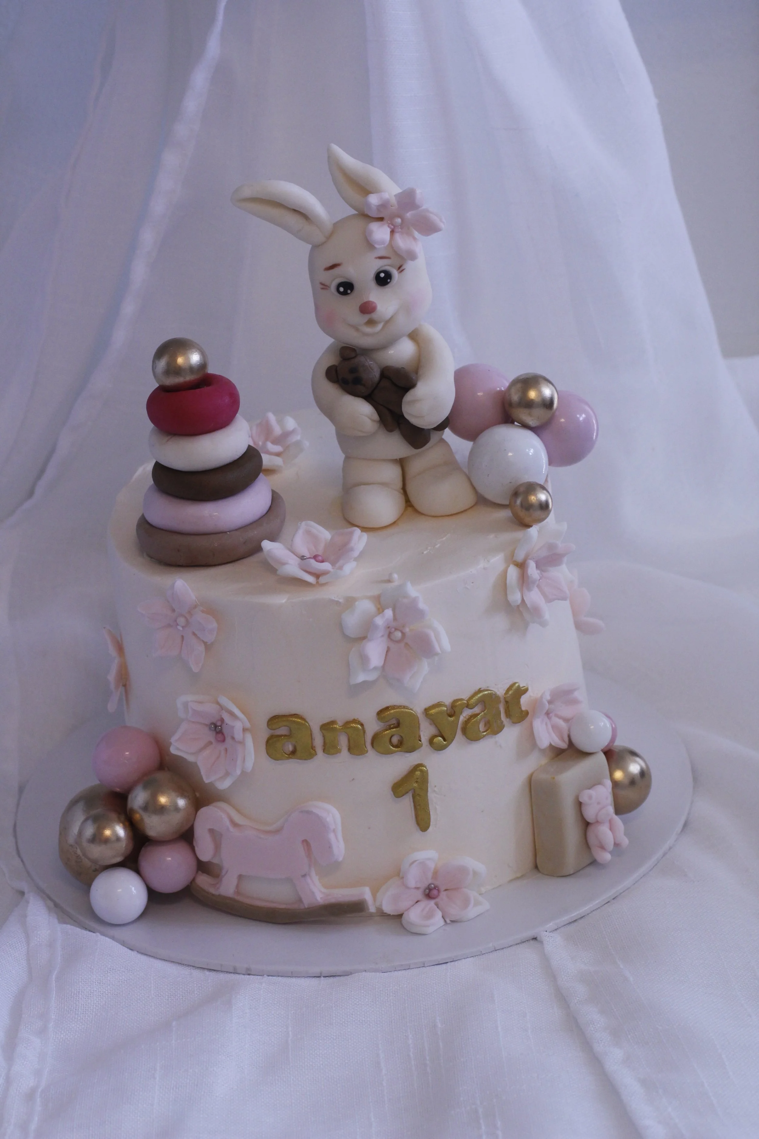 A pastel-colored baptism cake decorated with bunny and floral themes, with the name 'Anayat' and the number 1 written on it.