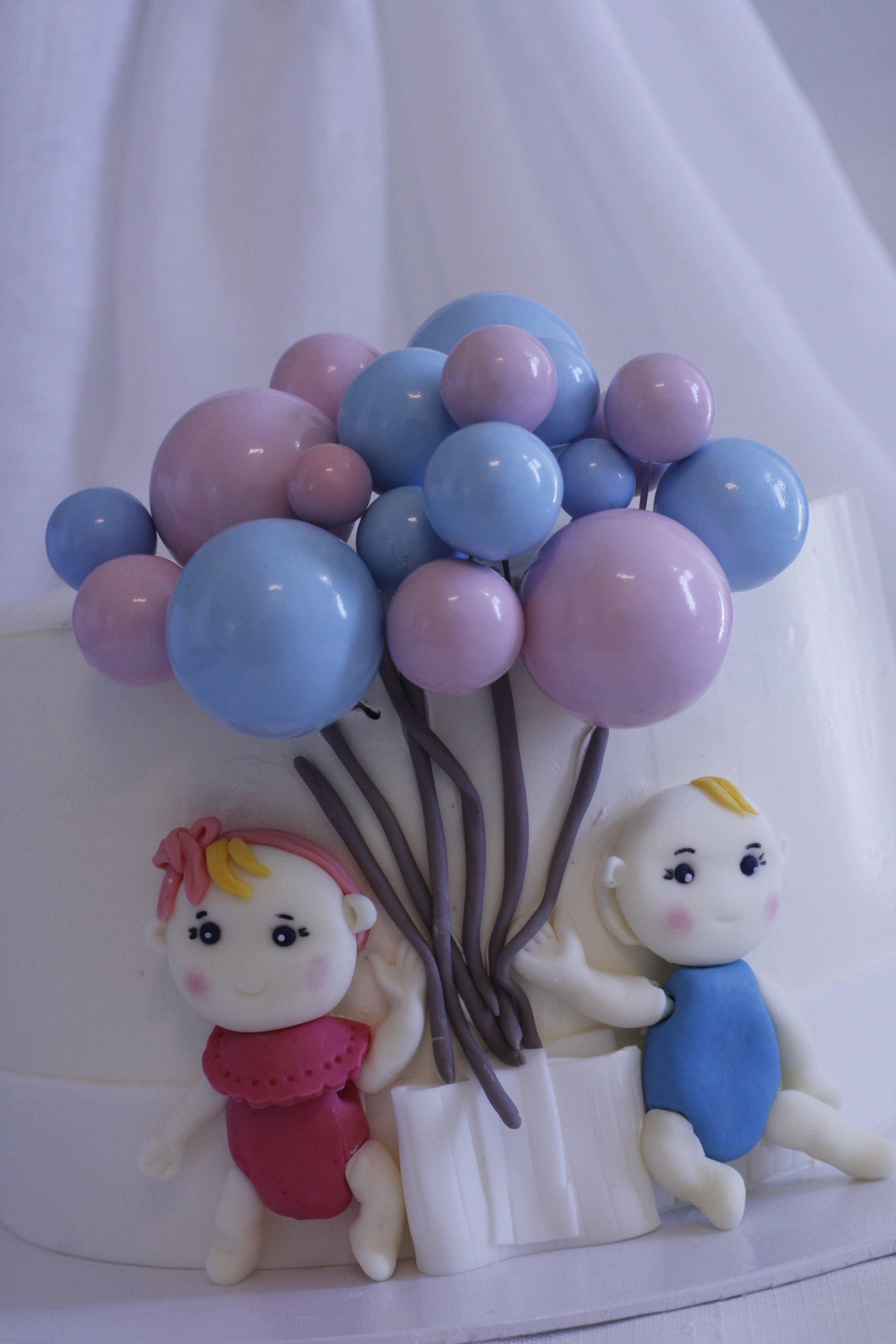 Decorative cake featuring two fondant children sitting beside a fondant balloon bouquet with pastel blue and pink balloons.
