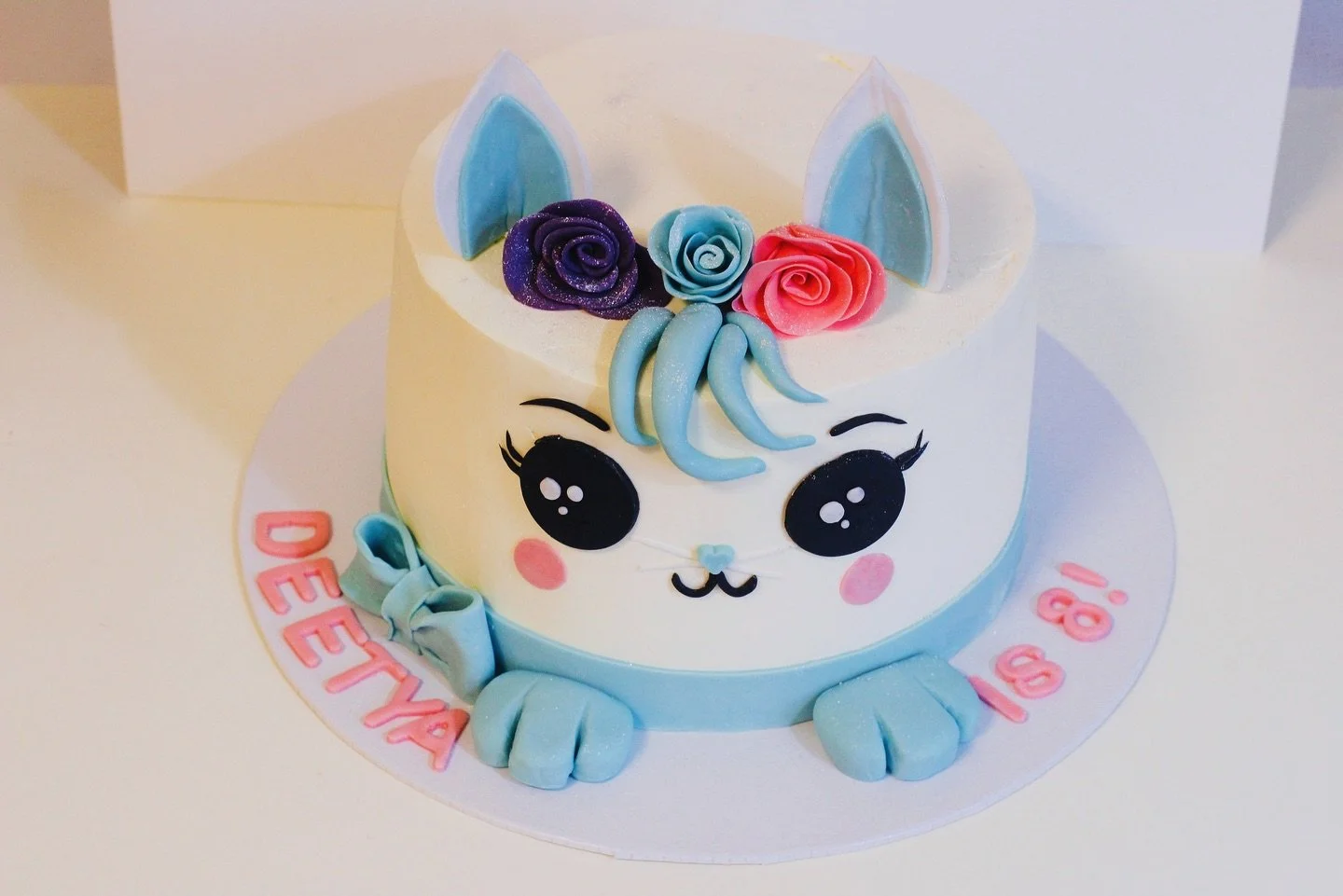 Unicorn-themed birthday cake with a cute face, colorful flowers, and pastel blue and pink accents, on a white surface.
