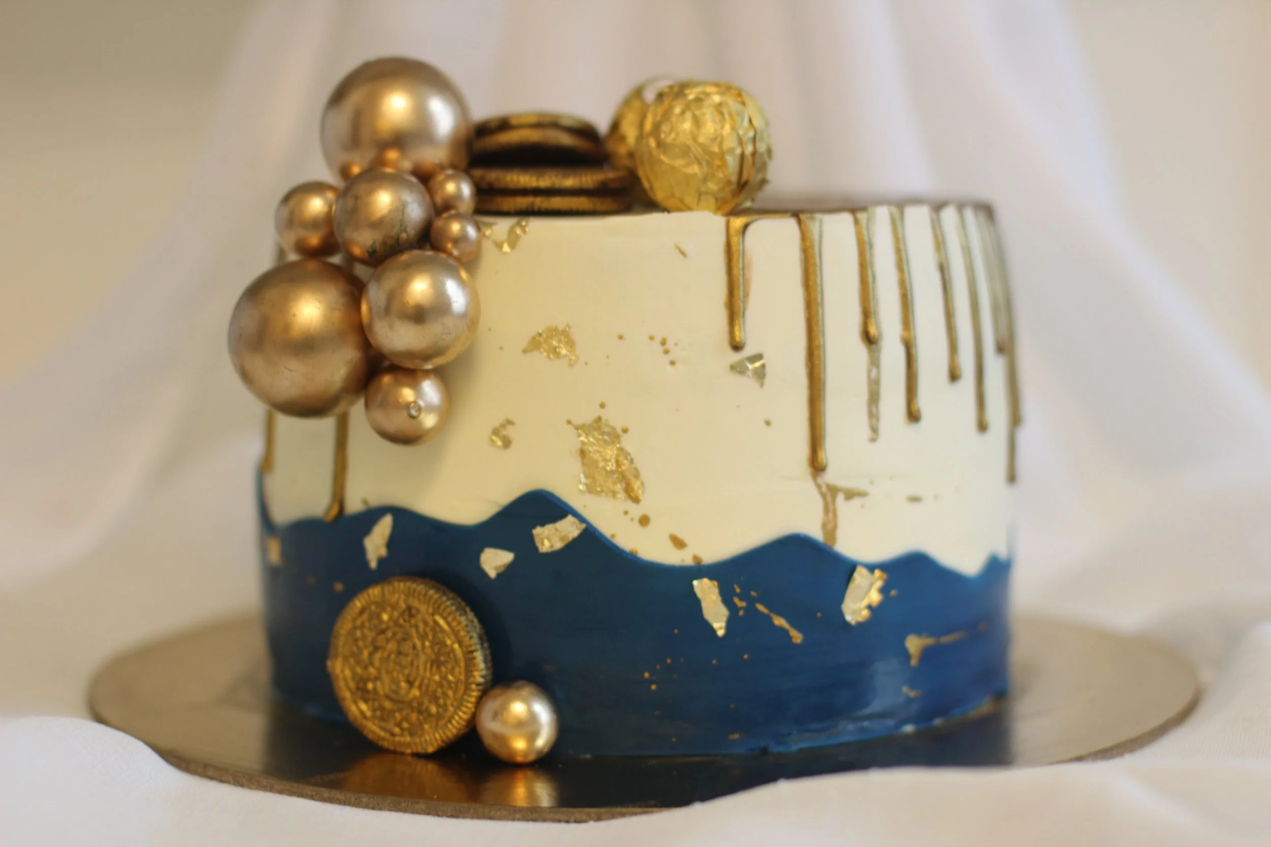 A decorated cake with gold and navy blue accents, topped with gold spheres and chocolate coins, on a round golden base.
