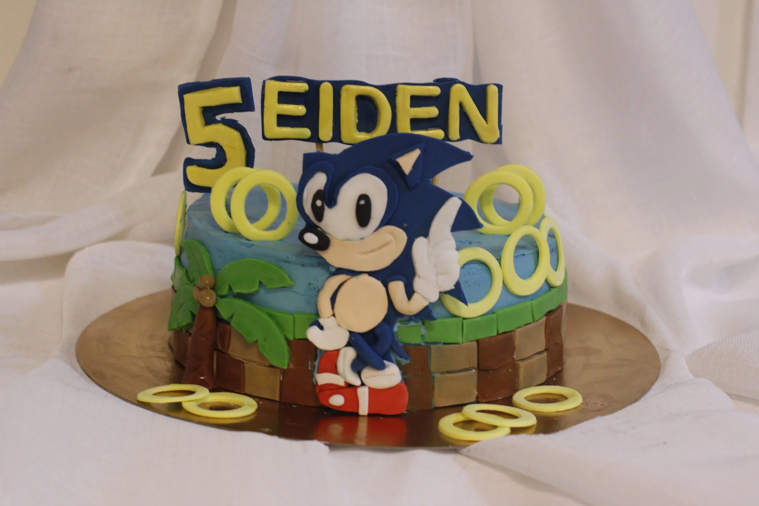 Cake decorated with Sonic the Hedgehog, palm trees, and yellow rings, with a sign that reads '5 EIDEN' on top, celebrating a fifth birthday.