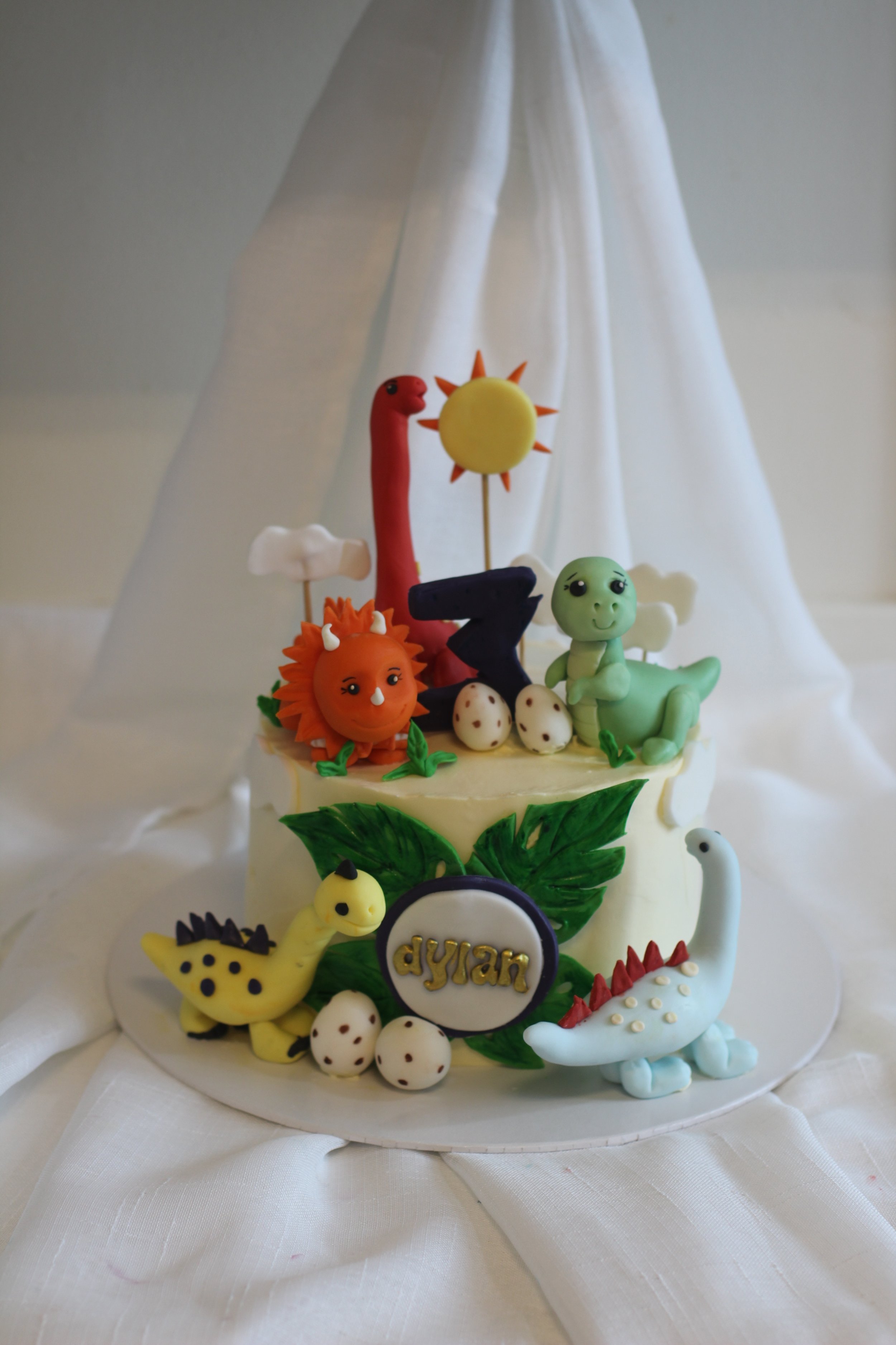 Colorful dinosaur-themed birthday cake with various dinosaur figurines, a sun, a number 7, and a birthday name plaque on white cloth background.