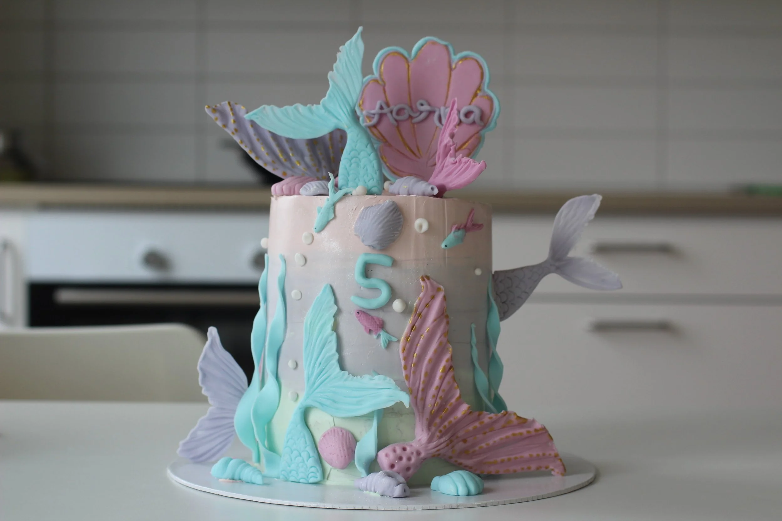 A mermaid-themed birthday cake decorated with pastel-colored mermaid tails, seashells, and fish, with the number 5 and a pink shell with the name 'Aarya' on top.