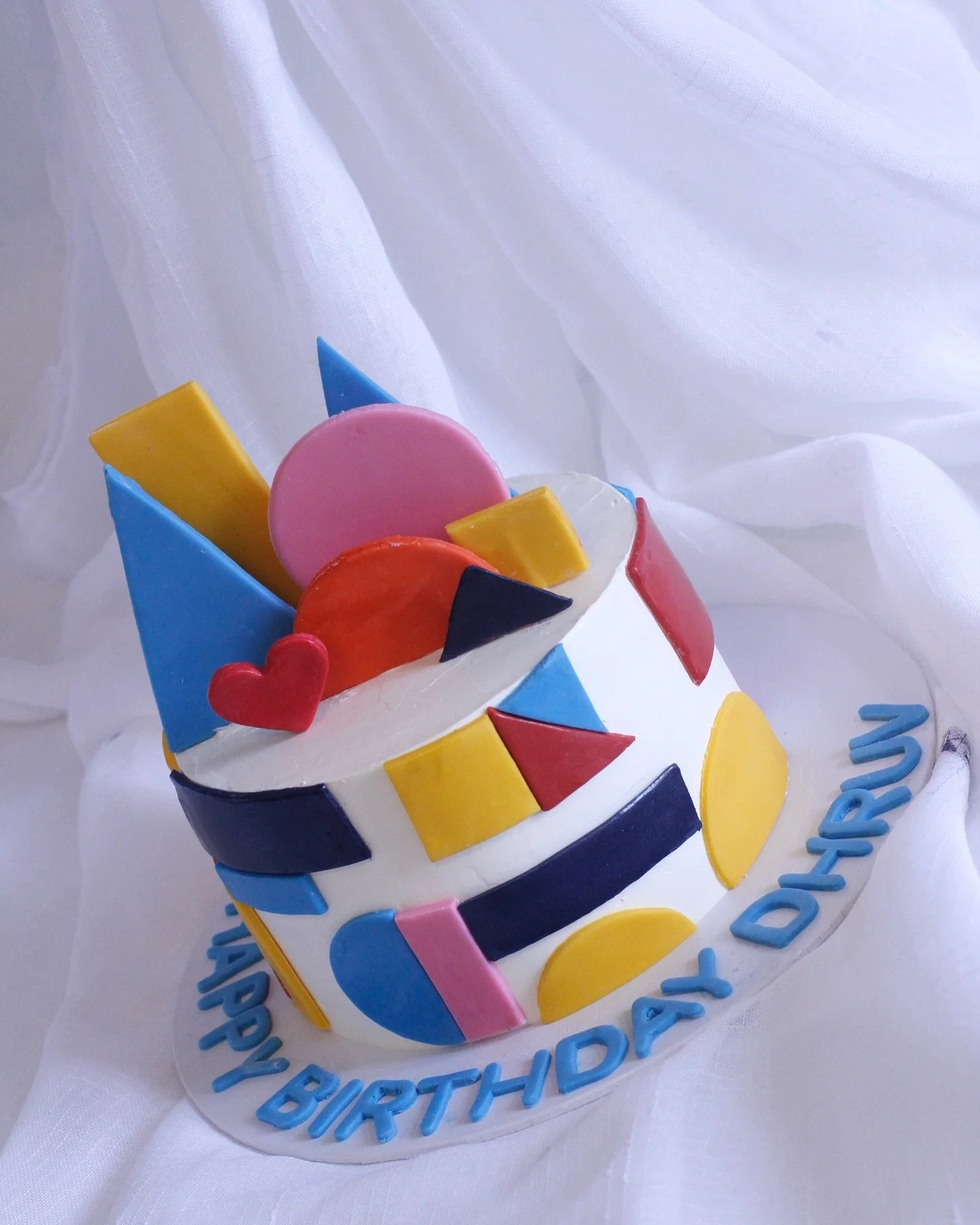 Colorful birthday cake decorated with geometric shapes and candies on top, with a 'Happy Birthday' message around the base, set on a white cloth background.