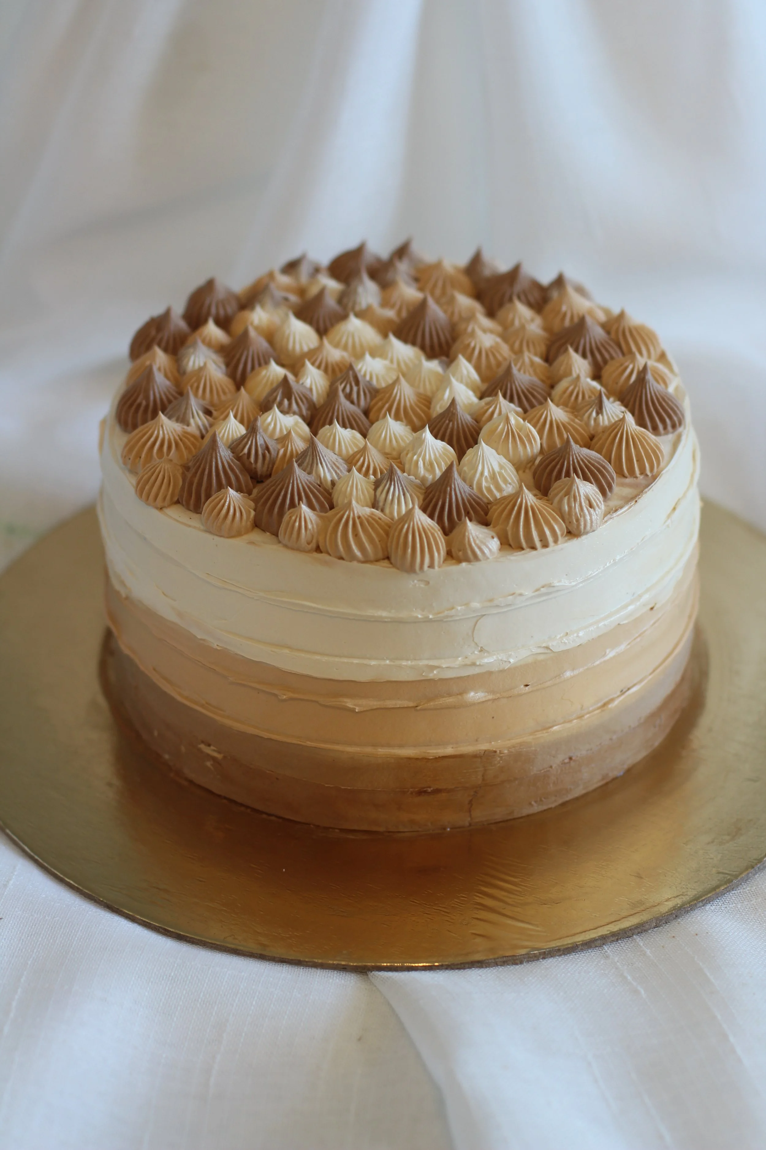 A layered cake with beige, white, and light brown frosting topped with dollops of frosting in similar shades, on a gold cake board.