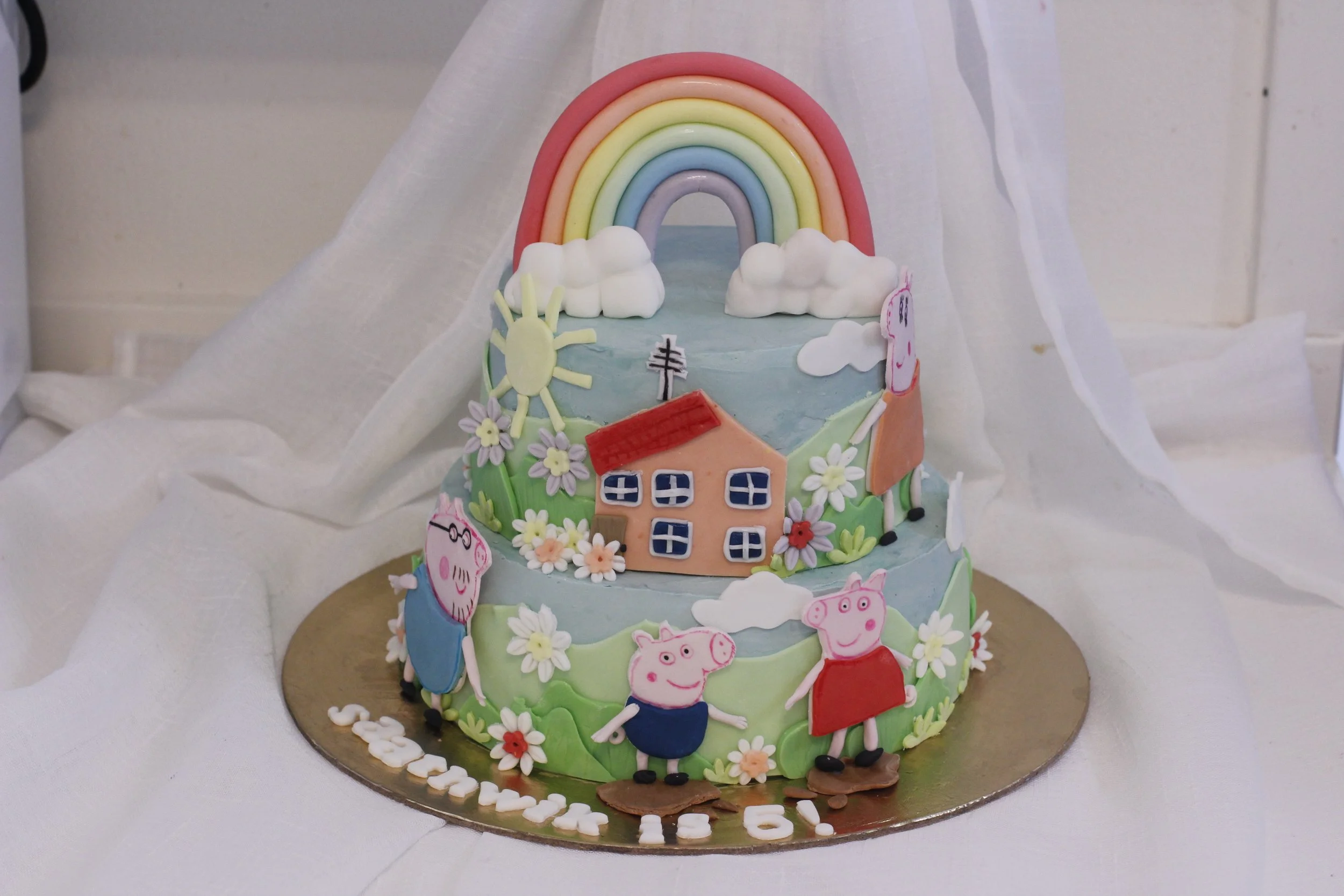 A tiered cake decorated with Peppa Pig characters, flowers, a house, a rainbow, clouds, a sun, and a figure of Miss Rabbit, celebrating a 6th birthday.