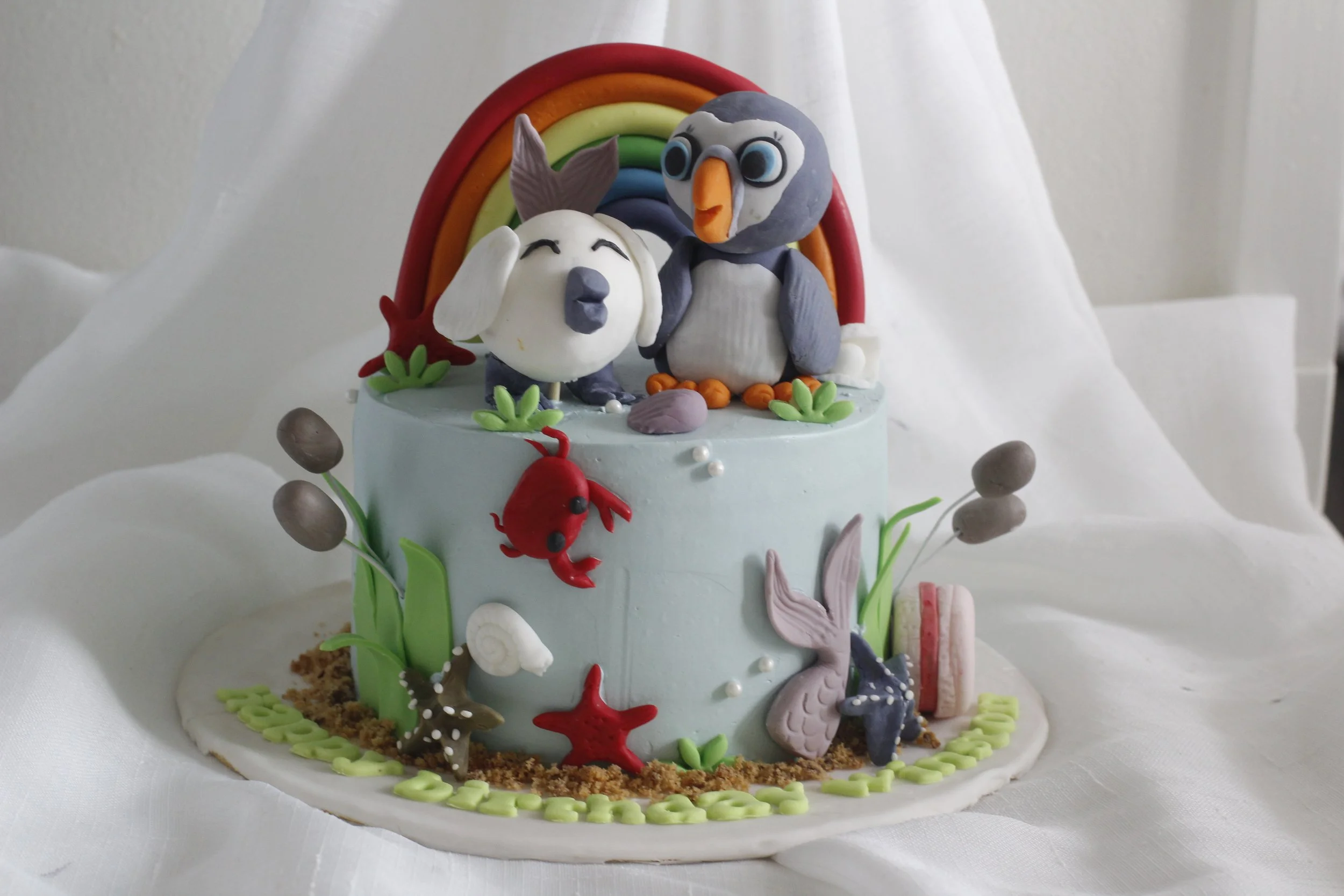 A beach-themed cake decorated with ocean animals and a rainbow, featuring a dog and a penguin figurine on top.