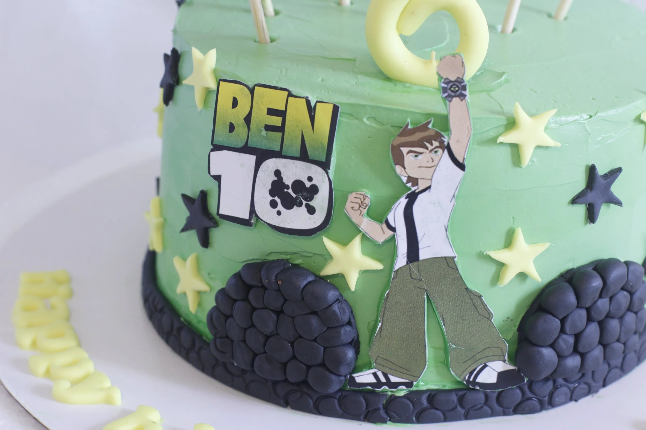 A green birthday cake decorated with cartoon characters, stars, and stones, celebrating Ben 10's 10th birthday.