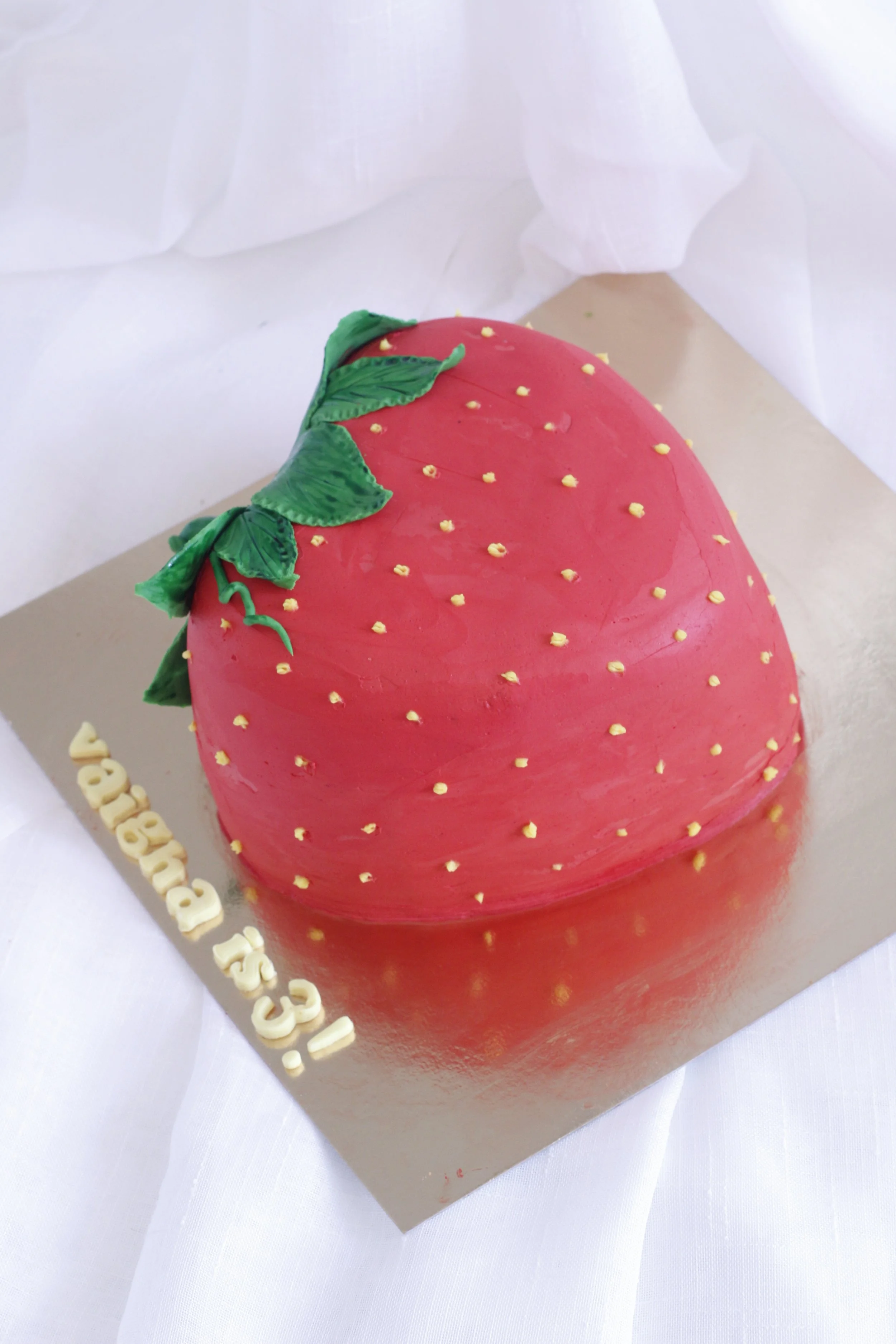 Chocolate cake shaped like a large strawberry with green leaves on top, decorated with yellow sprinkles, on a gold-colored base with 'Happy 45!' written on it.
