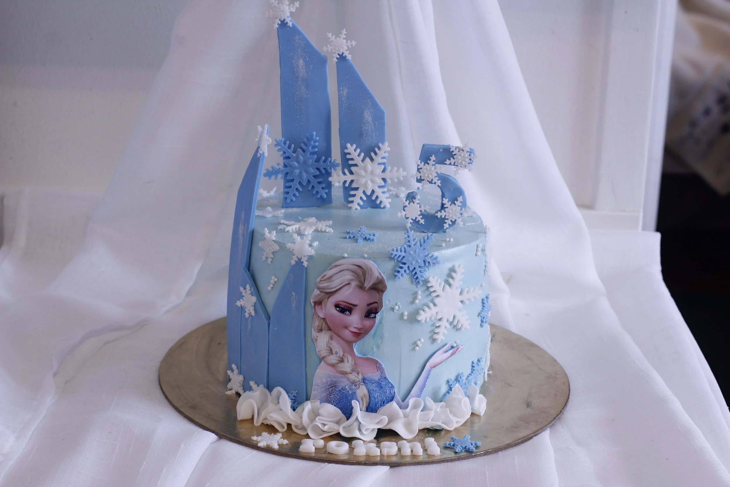 A Frozen-themed birthday cake with Elsa's image, blue and white snowflake decorations, and a number 5 topper, placed on a round gold cake board with white frosting accents.