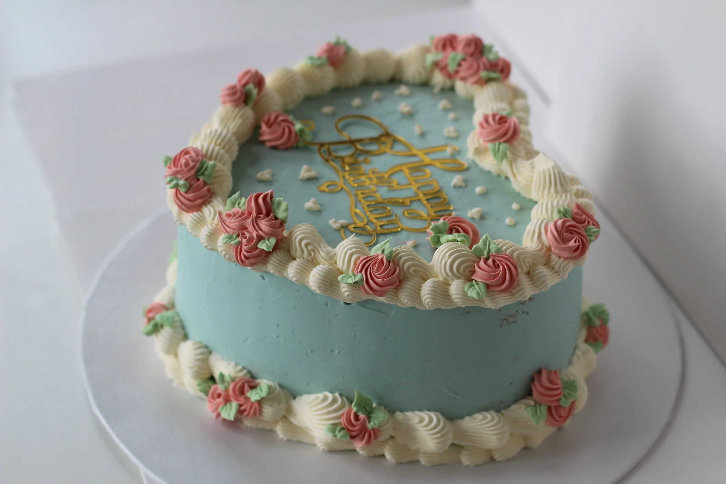 A heart-shaped cake decorated with blue frosting, pink rose-like icing flowers, white piping borders, and a gold message on top.