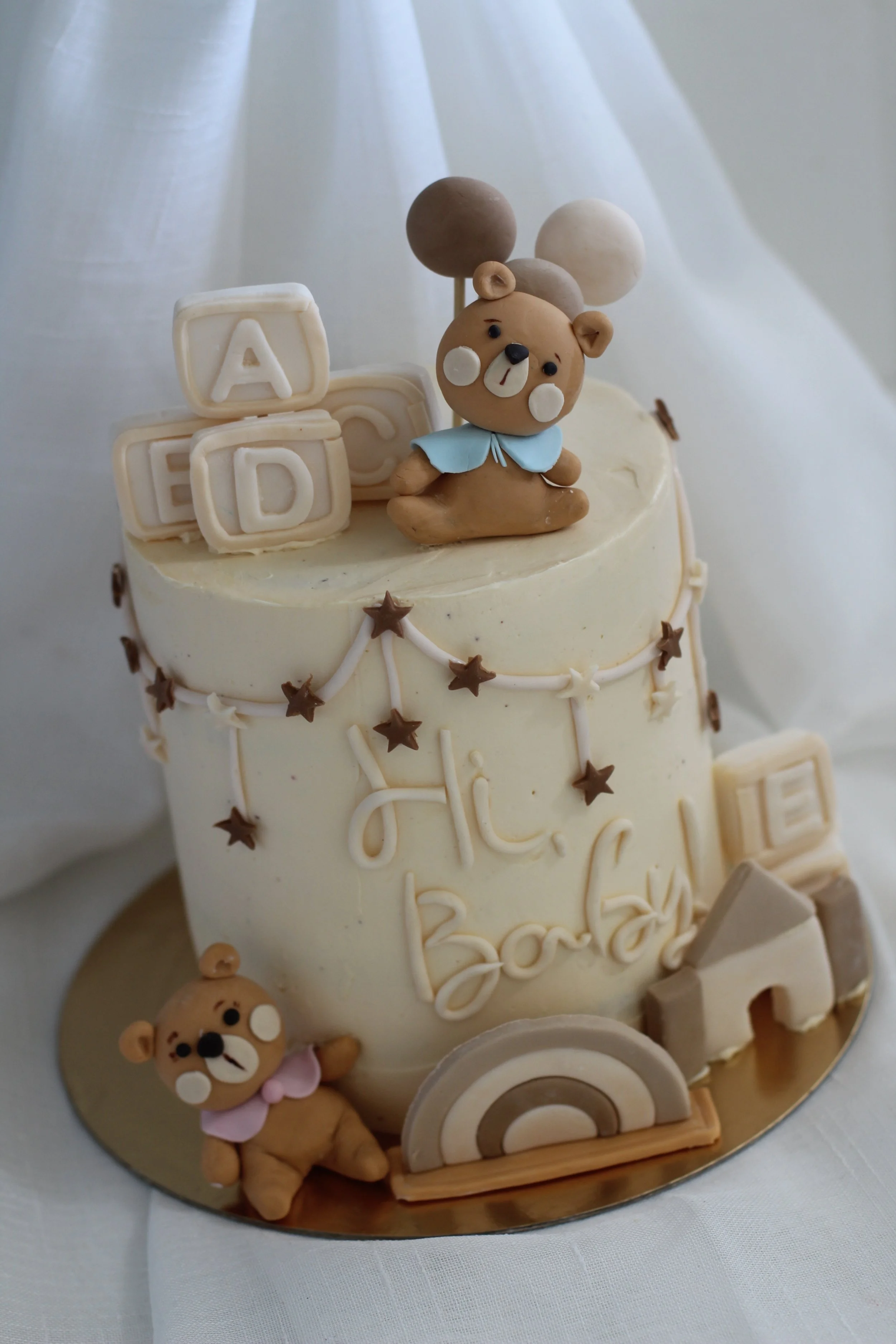 Cream-colored birthday cake with teddy bear and alphabet block decorations, featuring a teddy bear figurine on top, rainbow, stars, and the words "Hi Baby" written on the front.
