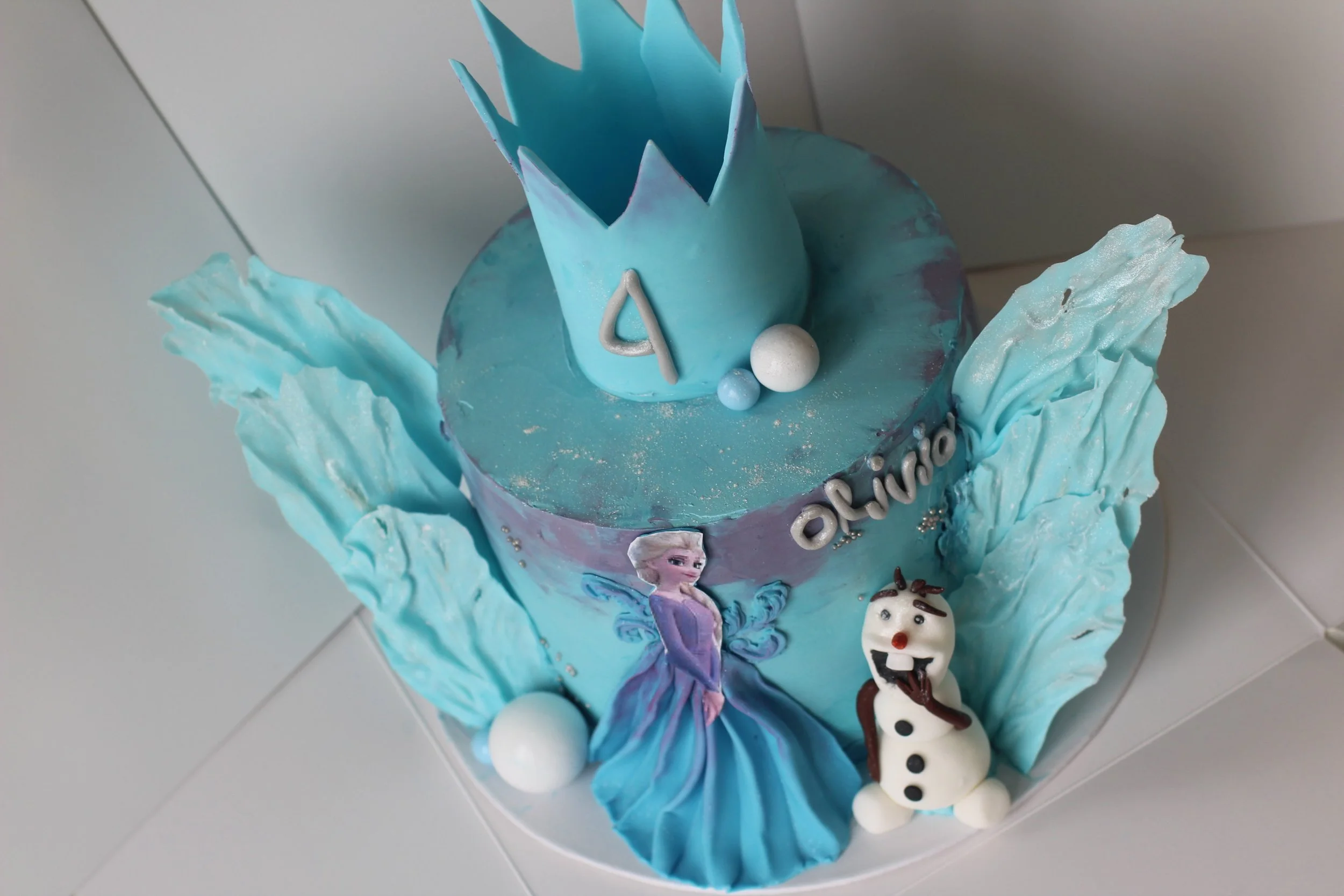 A birthday cake themed from Frozen, with characters Elsa and Olaf, decorated in blue shades, with a crown and snowflake designs.