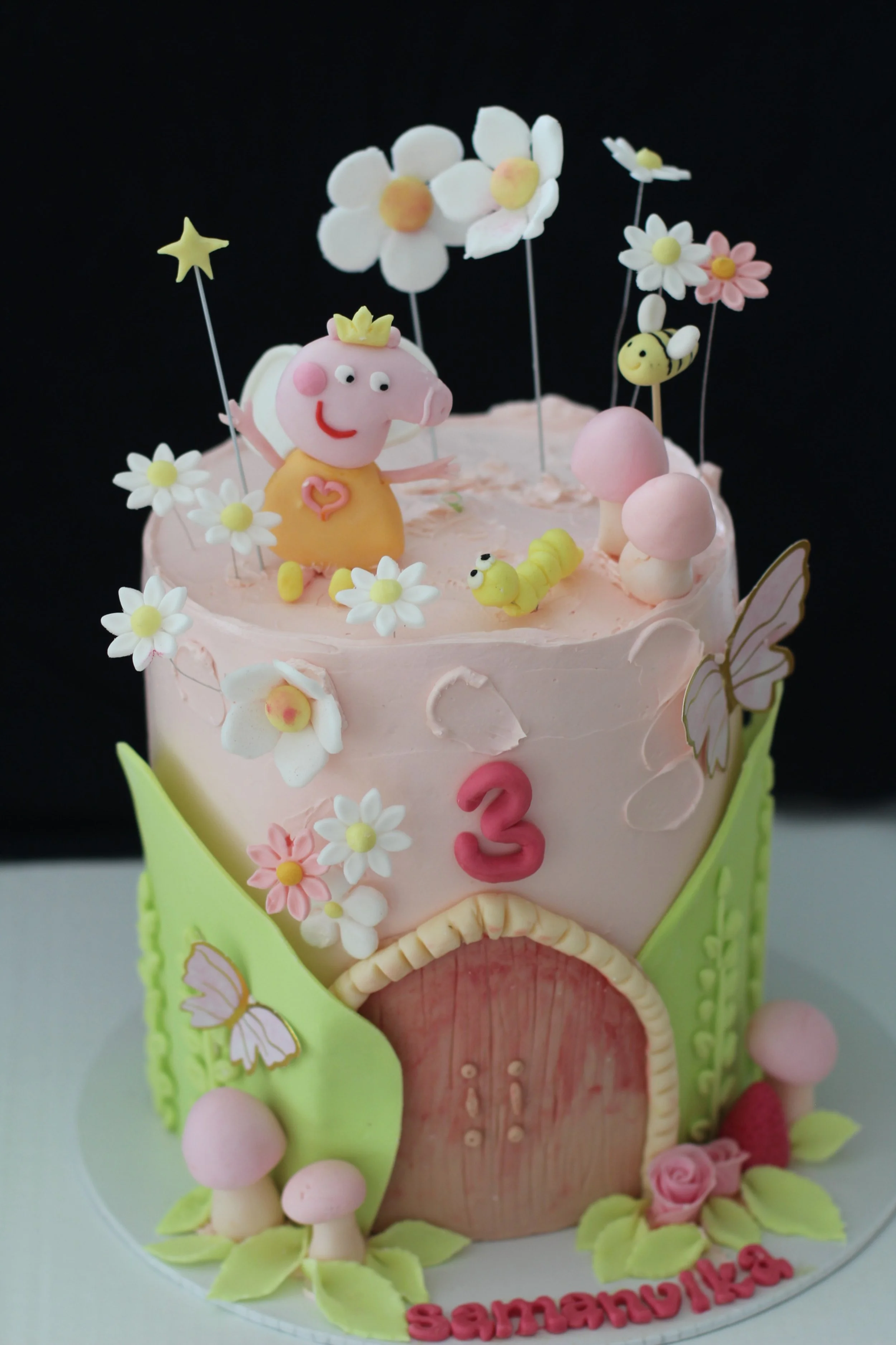 A pink and green birthday cake decorated with flowers, butterflies, mushrooms, a pig figure with a crown, and small caterpillars. The cake has a number 3 on it and a name "Samantha" at the bottom.