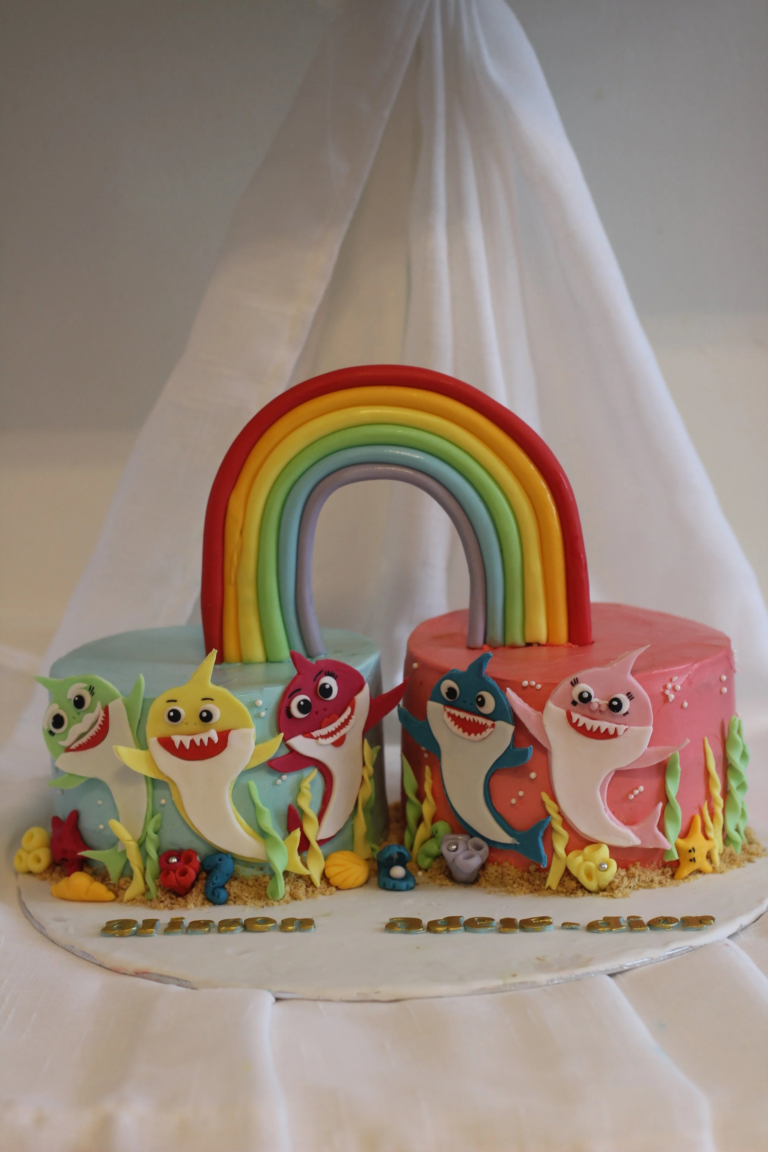 Colorful birthday cake with shark and dolphin decorations, a rainbow arch on top, and the words "birthday girl" written on the cake, set on a white tablecloth.