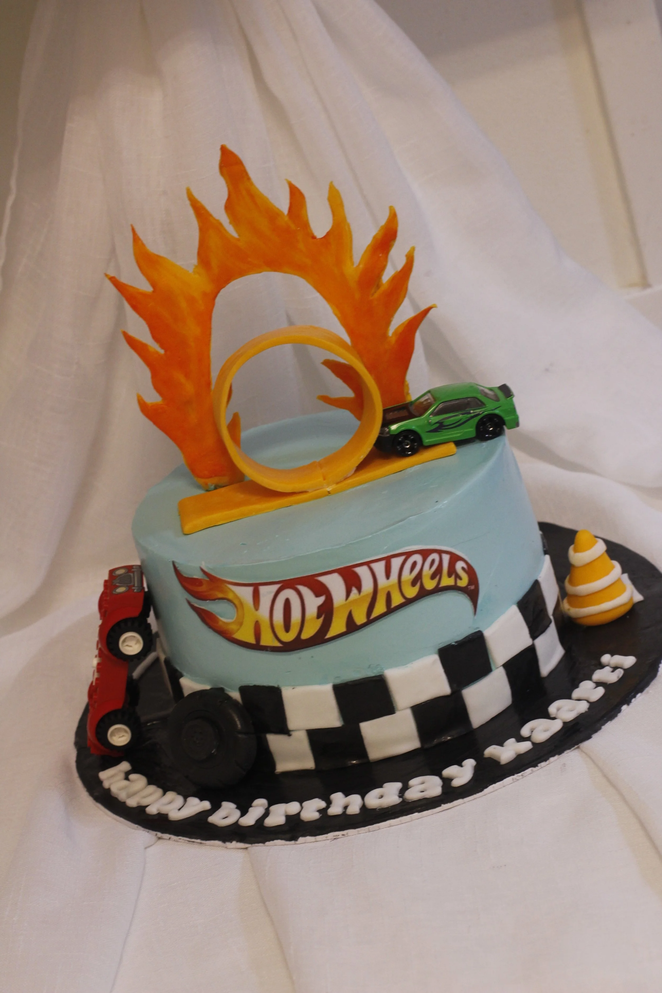 A birthday cake decorated with Hot Wheels theme, featuring a racing car, checkered flags, a fire-shaped decoration, and a small cone-shaped decoration, with the message 'happy birthday 12-20-22' on the base.