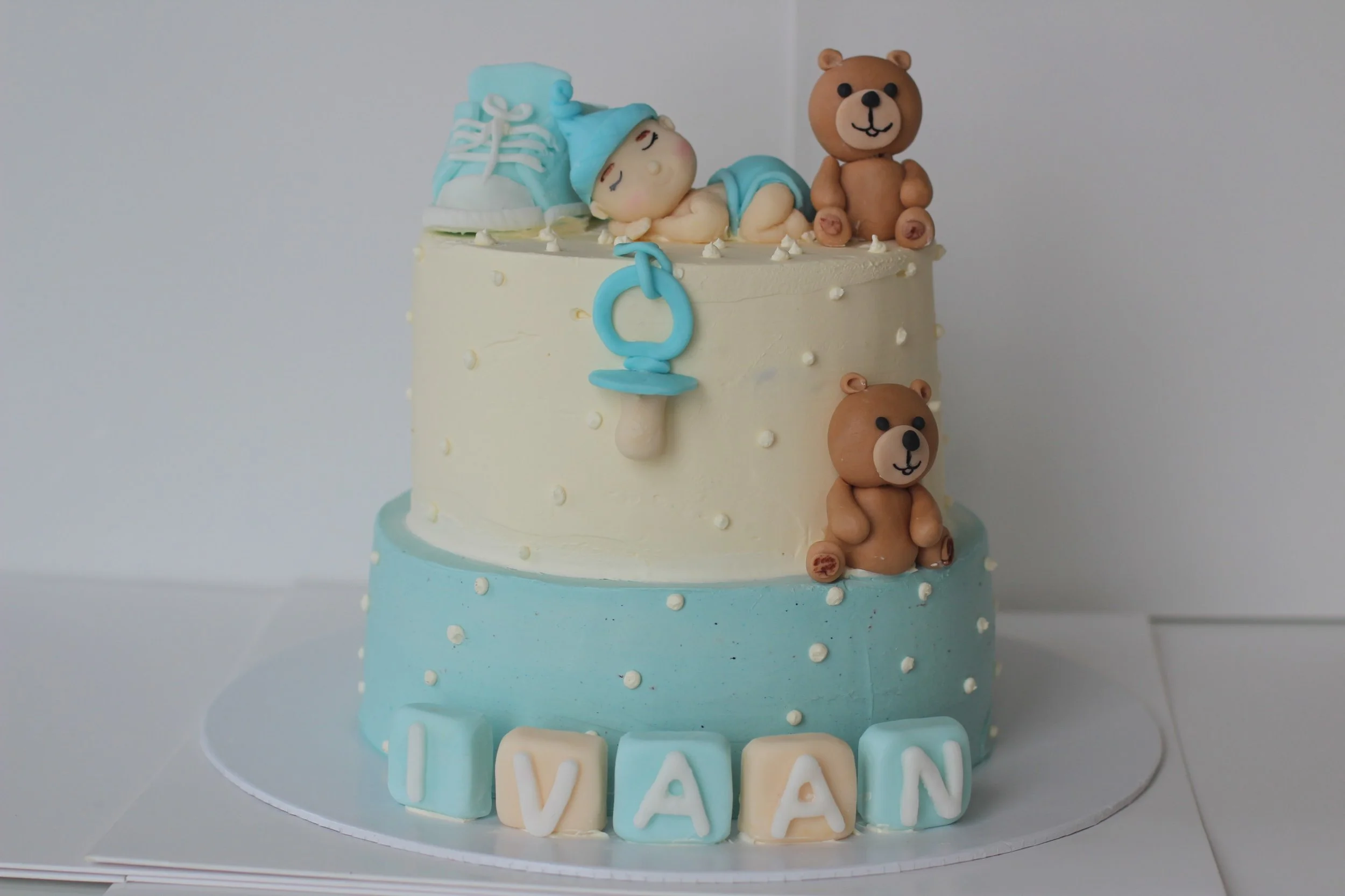 Two-tiered baby shower cake with blue and white frosting, topped with baby-themed decorations including a fondant baby doll, teddy bears, a pacifier, and baby shoes. The cake spells out the name "IVAN" with pastel-colored letter blocks.