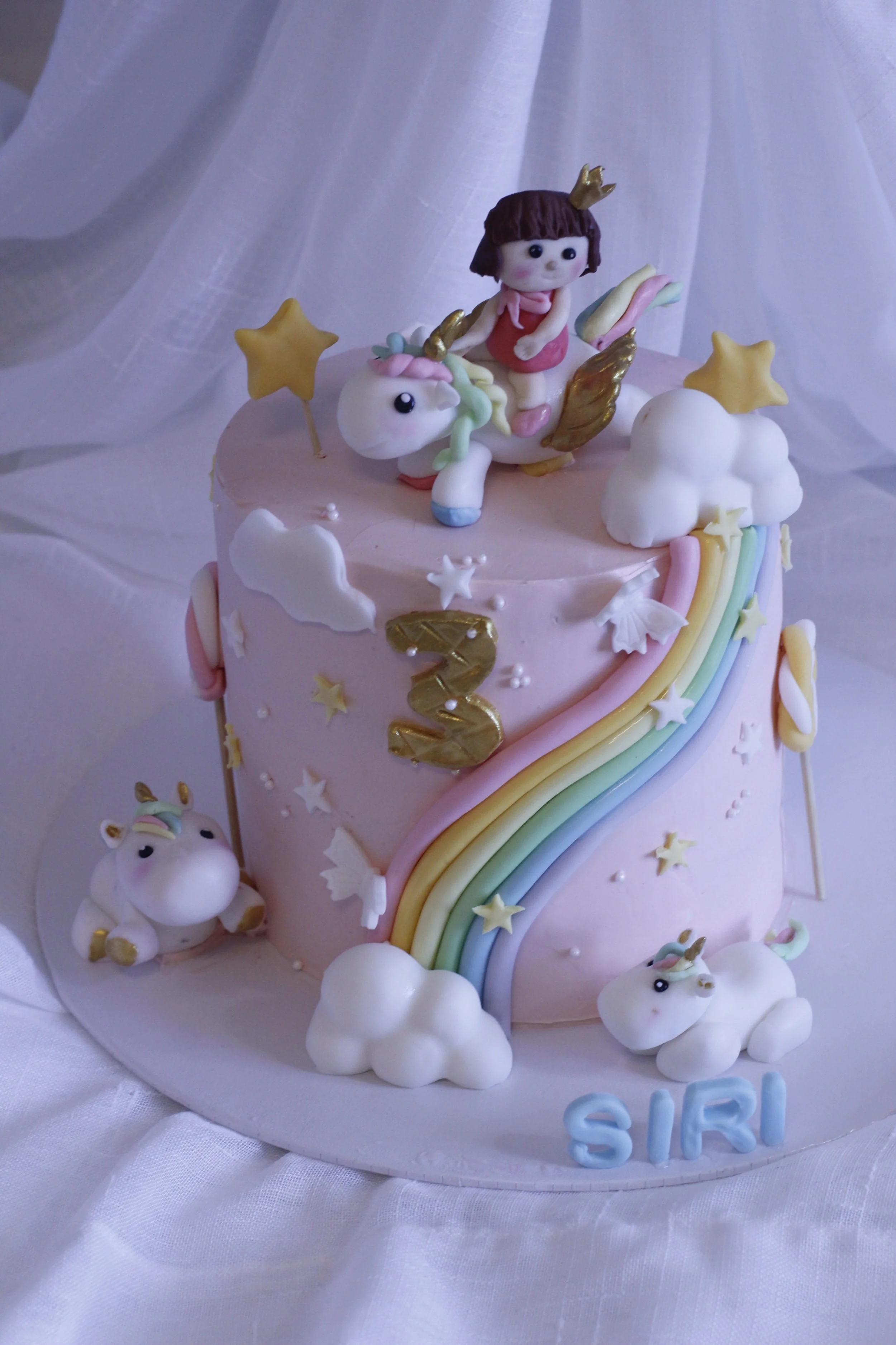 Pink birthday cake decorated with unicorns, rainbows, stars, clouds, and a girl figurine wearing a crown, celebrating a third birthday for Siri.