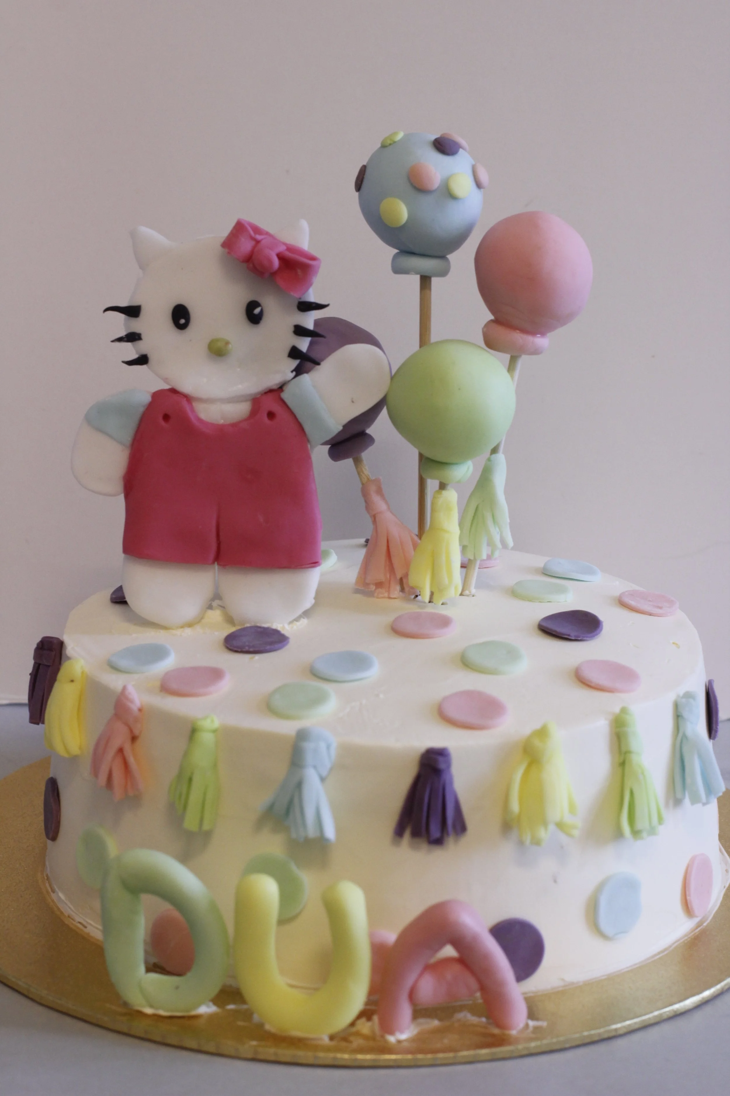 Colorful birthday cake with a Hello Kitty fondant figure wearing a pink dress and bow, surrounded by pastel-colored balloons and decorations.