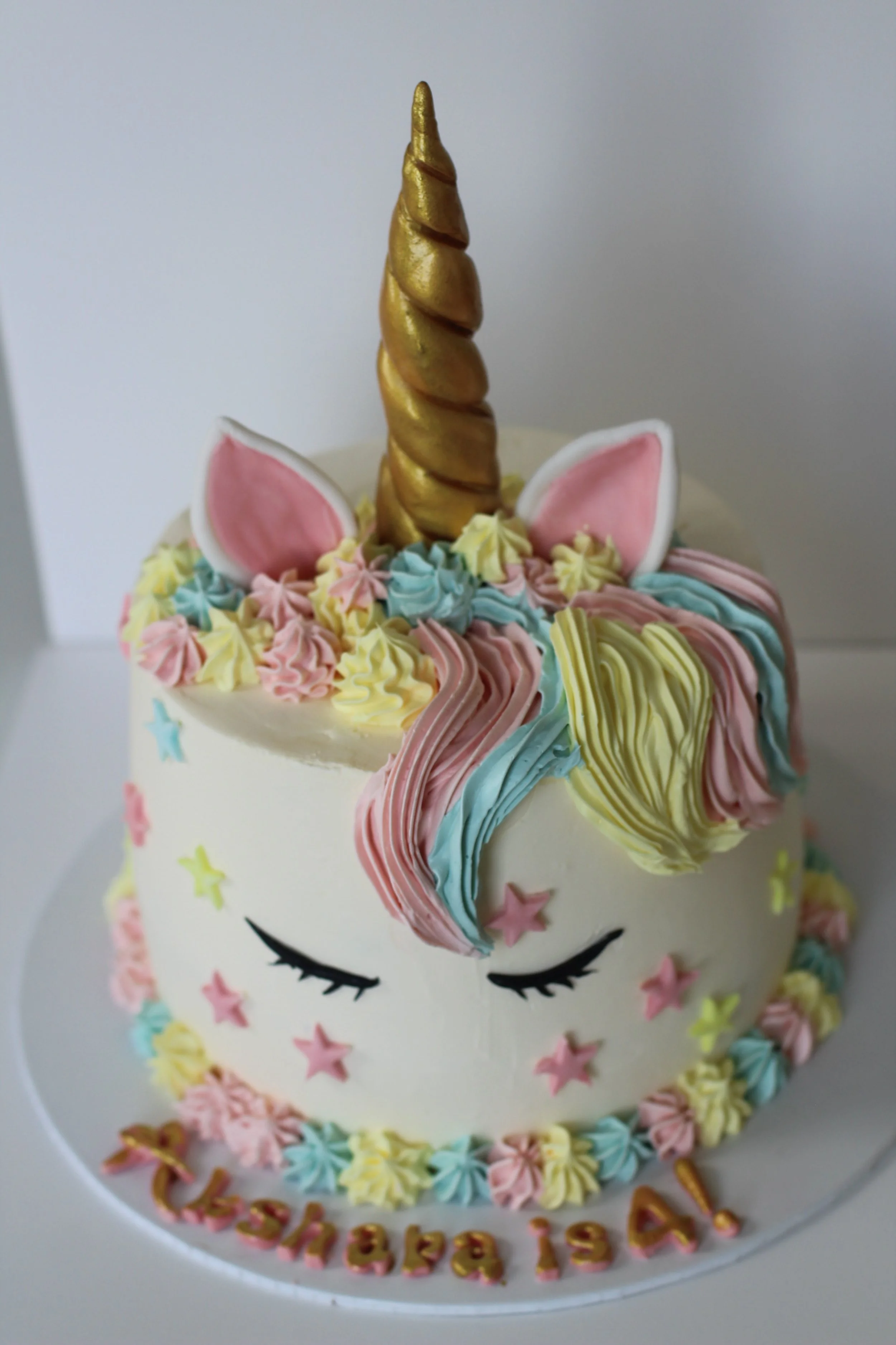 Unicorn-themed birthday cake with pastel-colored frosting, a gold horn, pink and white ears, closed eye decorations, and colorful star-shaped accents.