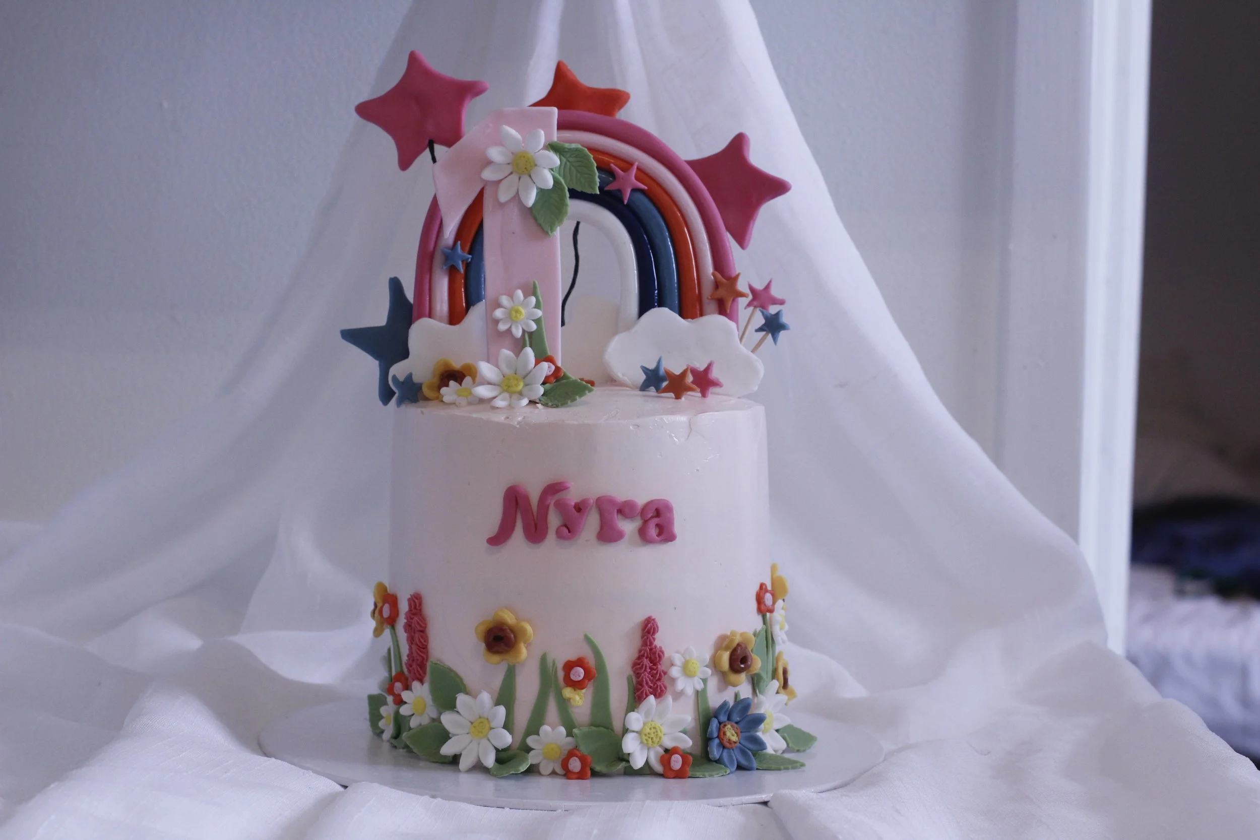 A pink birthday cake with colorful fondant decorations of a rainbow, stars, clouds, and flowers, with the name 'Nyra' written on the front.