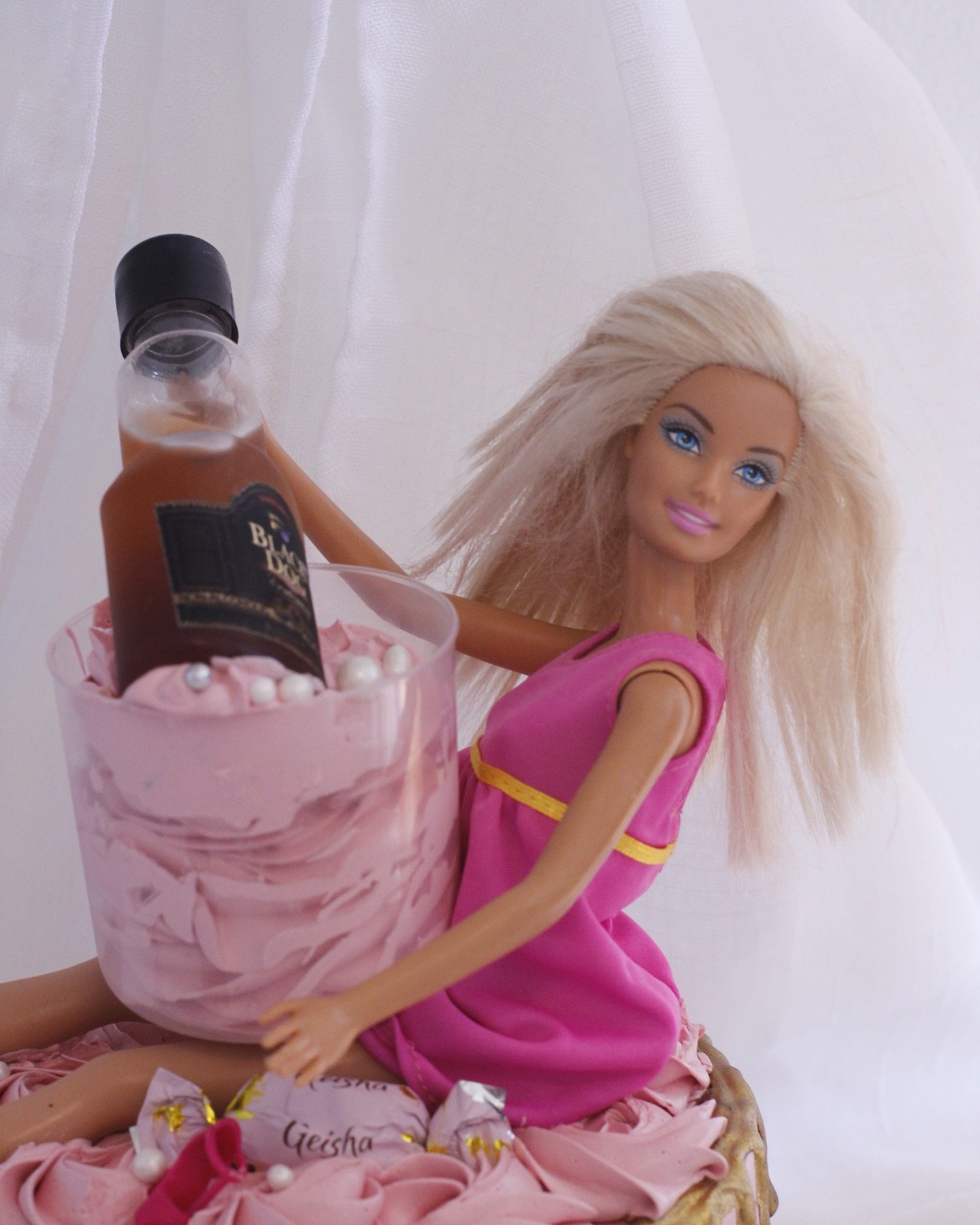 A Barbie doll with long blonde hair, wearing a pink dress, sitting on a pink birthday cake with white decorations, holding a large pink cup containing a bottle, against a white background.