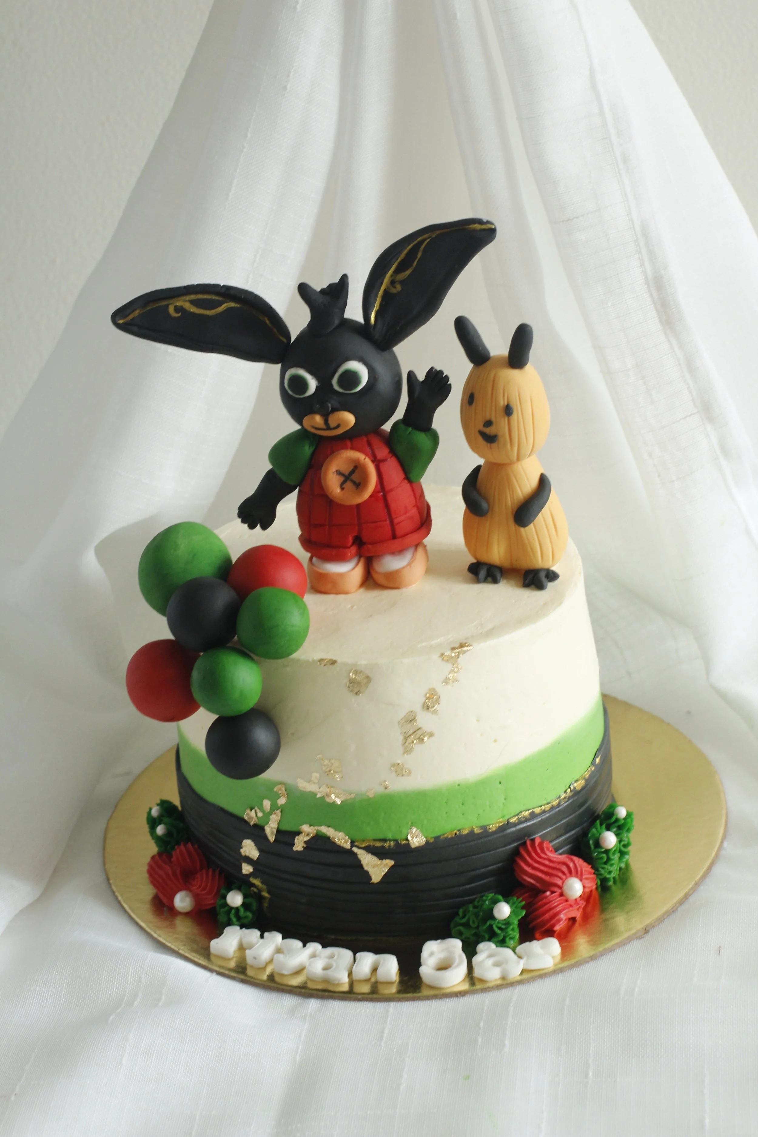 A decorated birthday cake with white, green, and black icing. The cake has a bunny and a smaller rabbit figure on top and colorful balloon decorations on the side. The base of the cake has the name 'Ivan' spelled out in white letters.