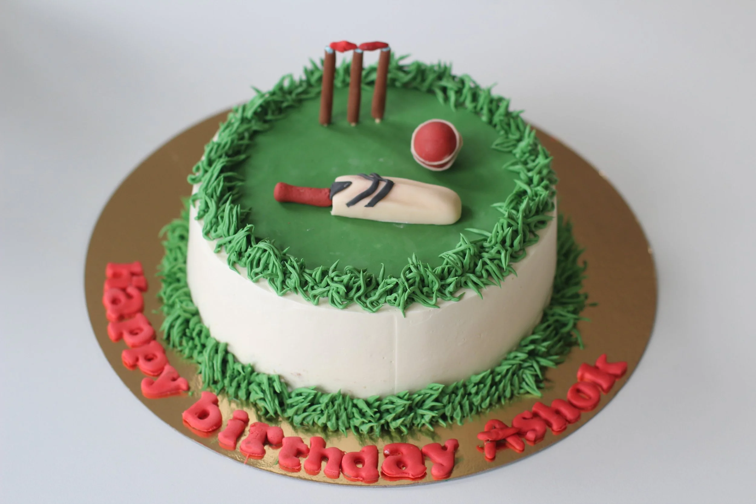 A round birthday cake decorated with a cricket field theme, including a white cricket bat, red ball, and three wickets, with green frosting resembling grass, and red lettering that says "Happy Birthday Tash" on a gold base.