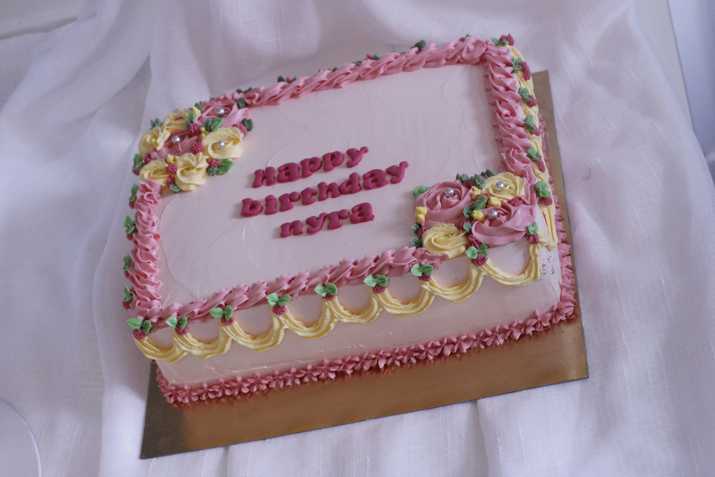 A rectangular birthday cake with pink icing, decorated with pink and yellow roses and green leaves, says 'Happy Birthday Nya' in pink frosting.