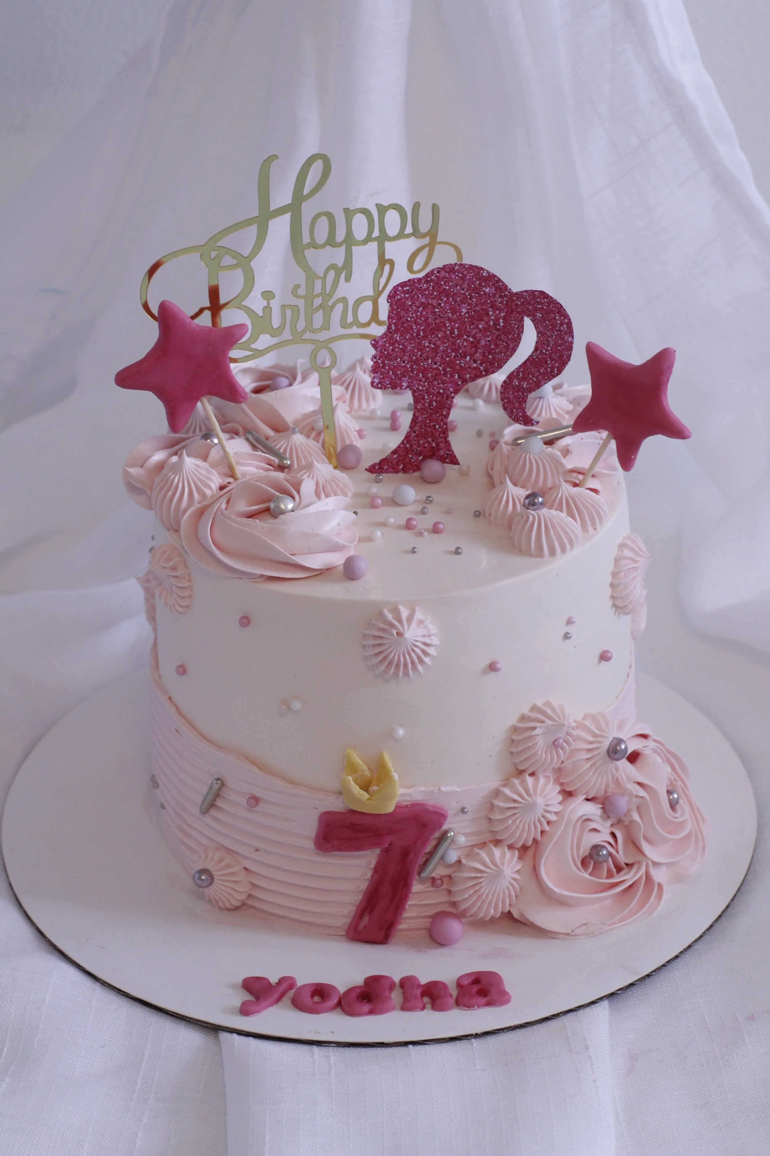 A two-tiered pink and white birthday cake decorated with pink roses, silver and pink pearls, and star-shaped toppers. The top tier features a pink glittery silhouette of a girl with a ponytail, a 