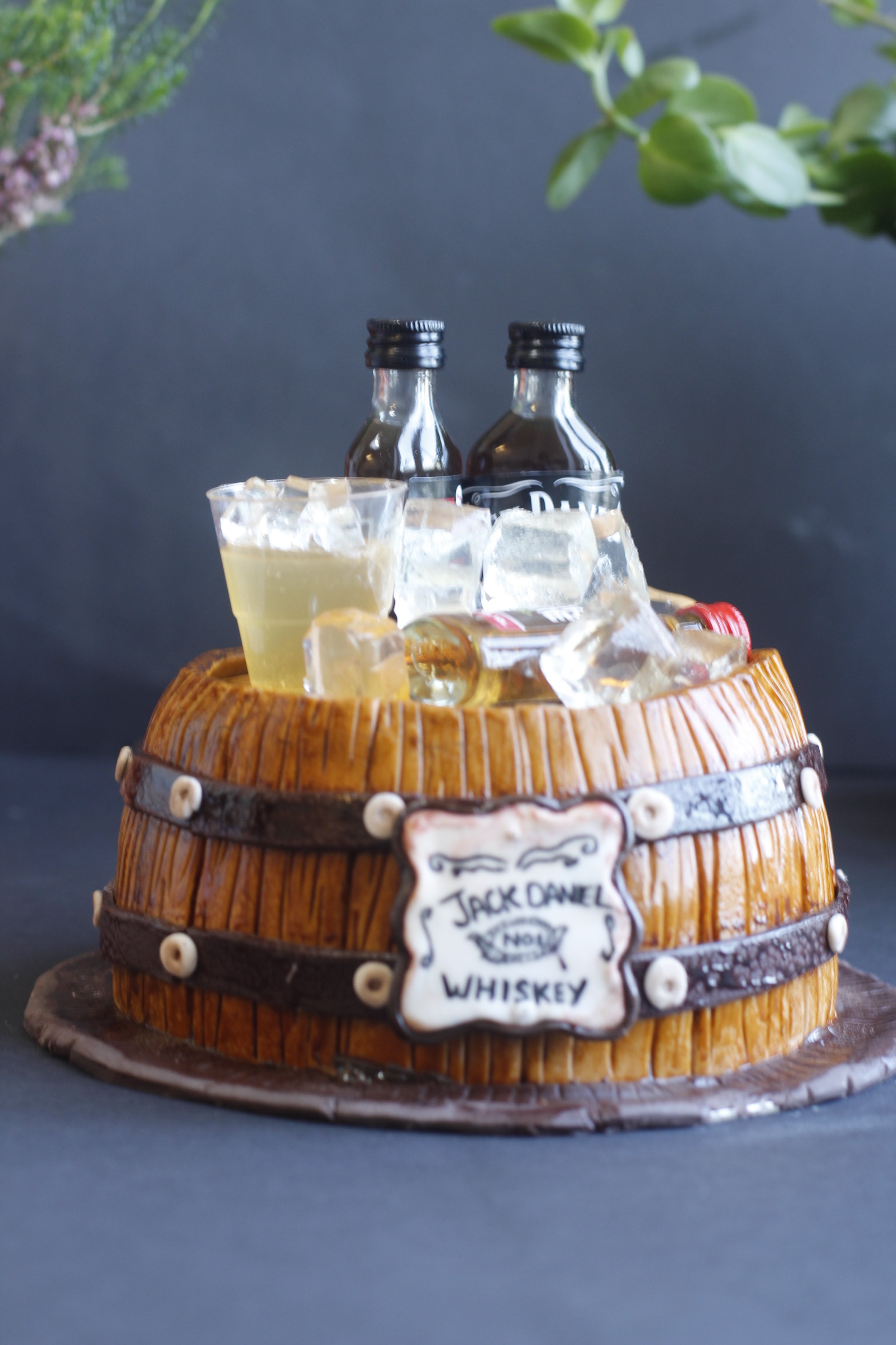 A cake designed to look like a whiskey barrel with bottles and glasses of whiskey on top.